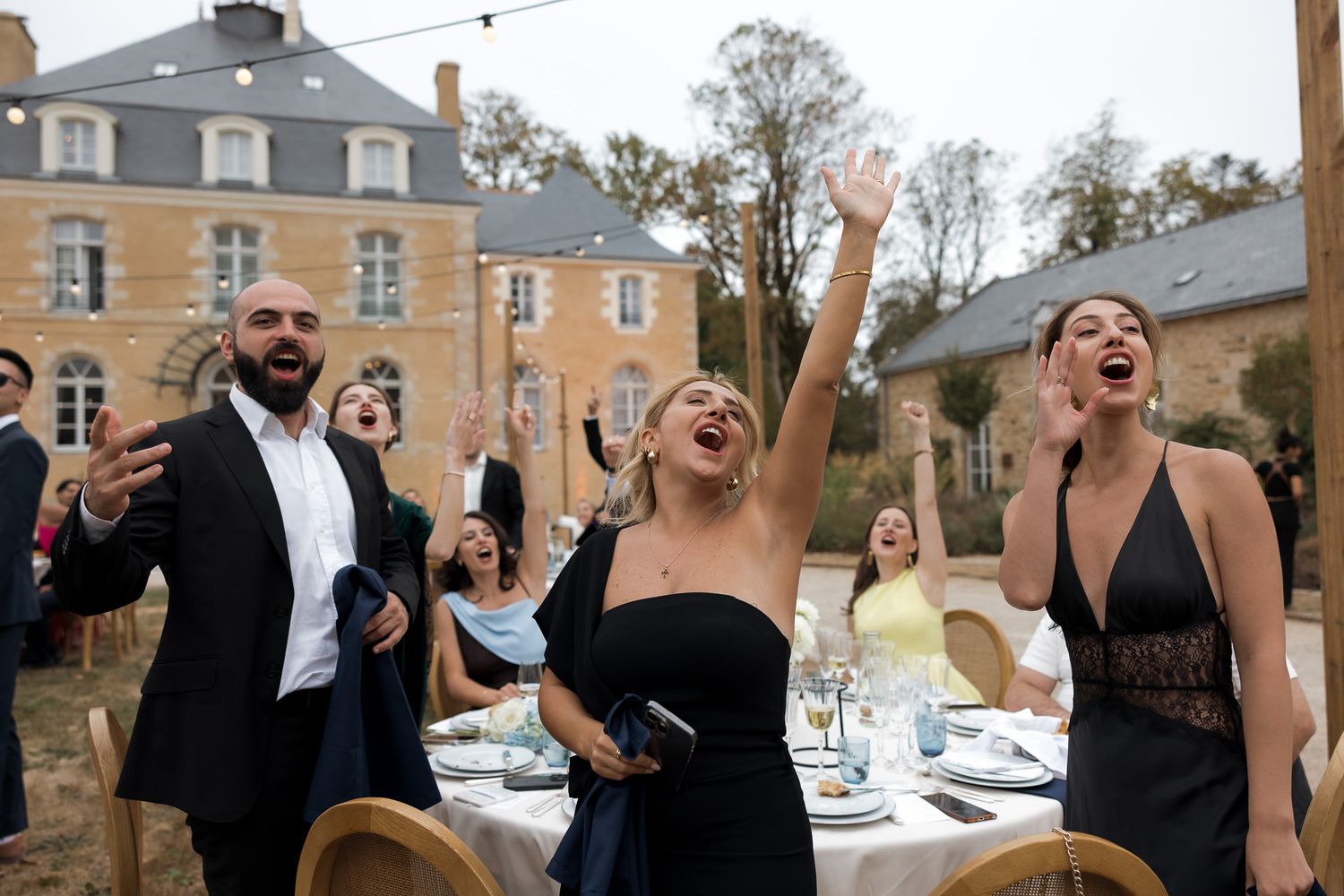 Joyful wedding guests celebrating at an outdoor reception in front of a French chateau, raising arms and singing.