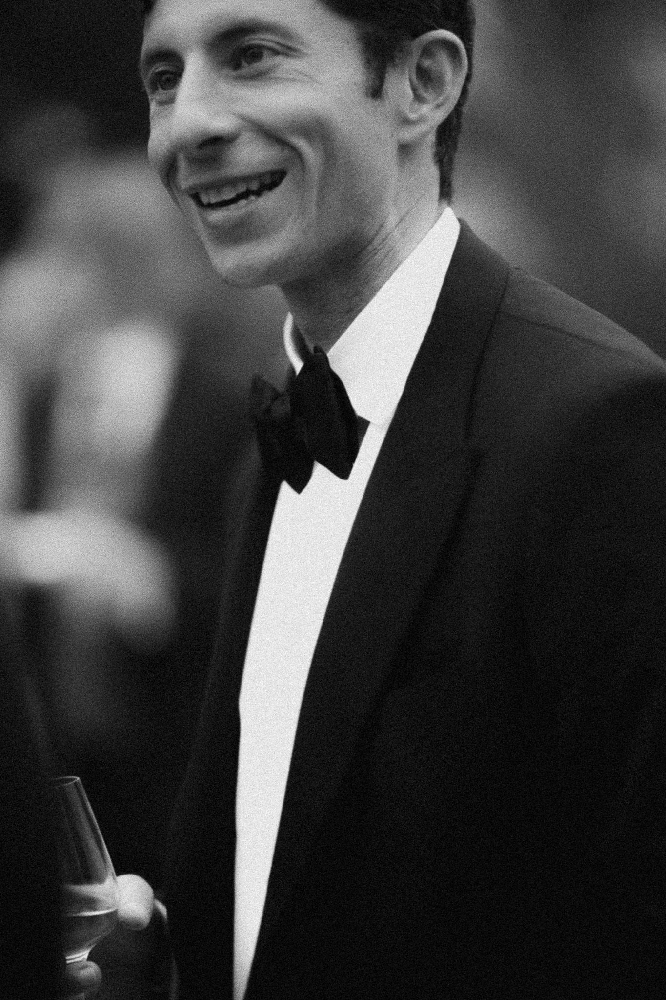 Smiling man in black tuxedo and bow tie holding a drink at a formal event, black and white photo.