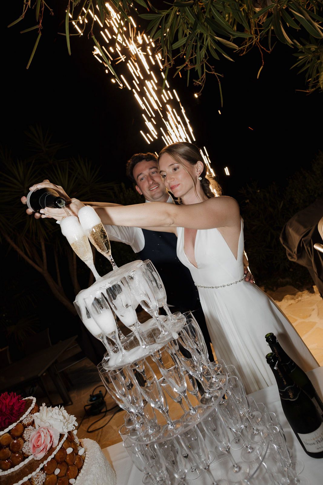 Couple pours champagne into tower of glasses with sparklers creating magical effect at wedding celebration.