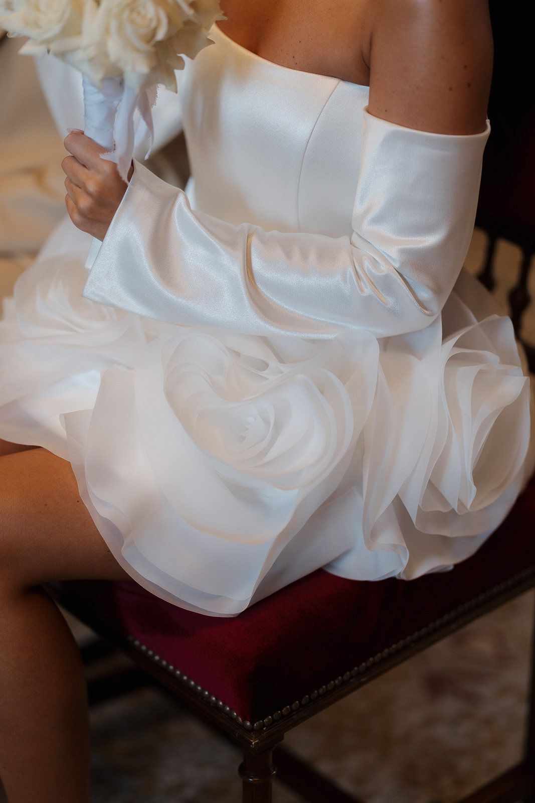 Close-up detail of white ruffled wedding dress fabric with layered organza roses sitting on red leather seat.