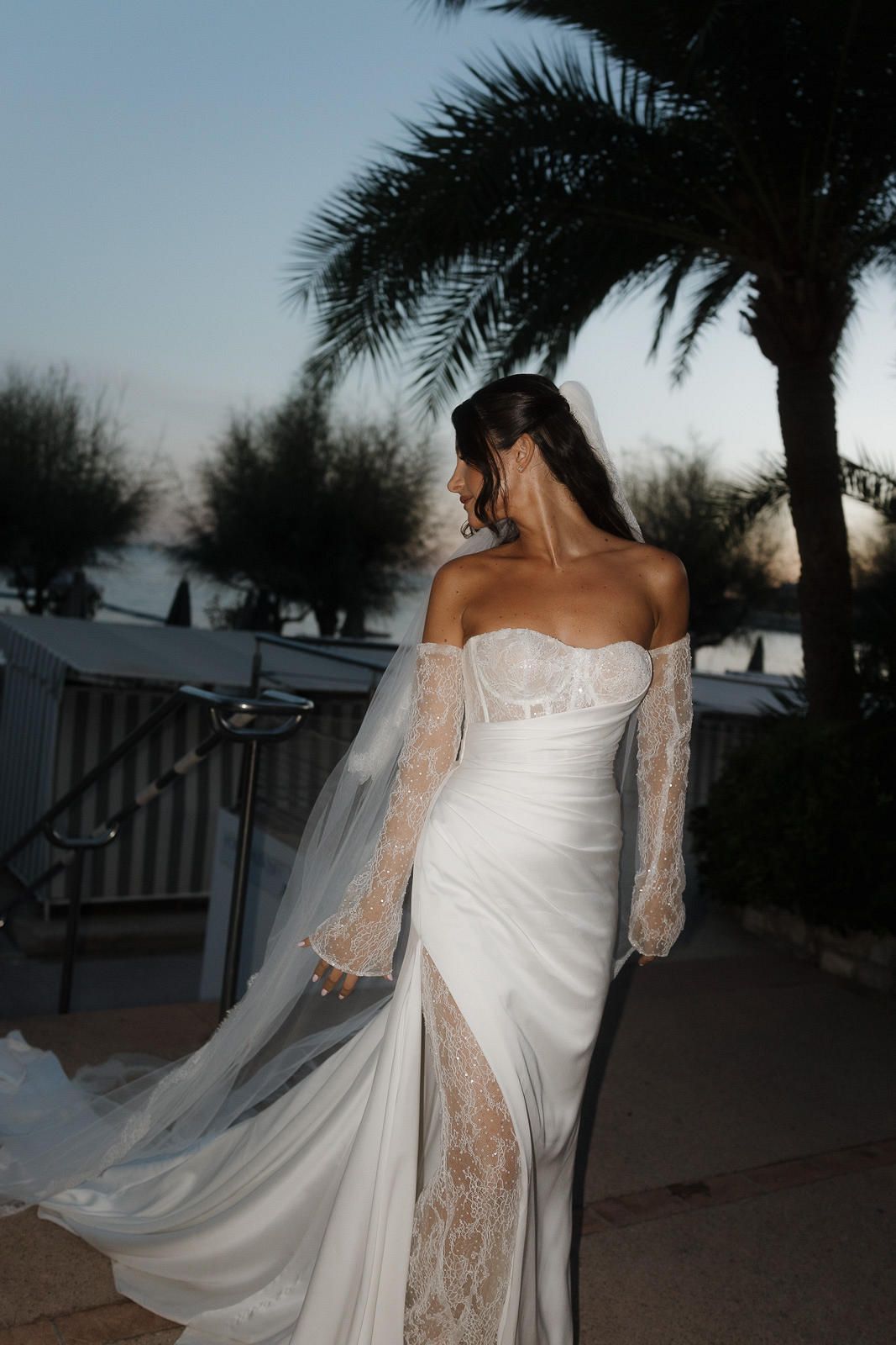 A bride in a flowing white wedding dress with lace sleeves poses at dusk under palm trees in a desert setting.