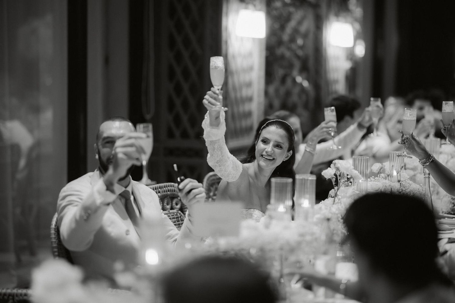 Black and white photo of wedding guests raising their glasses in a celebratory toast during a reception dinner.