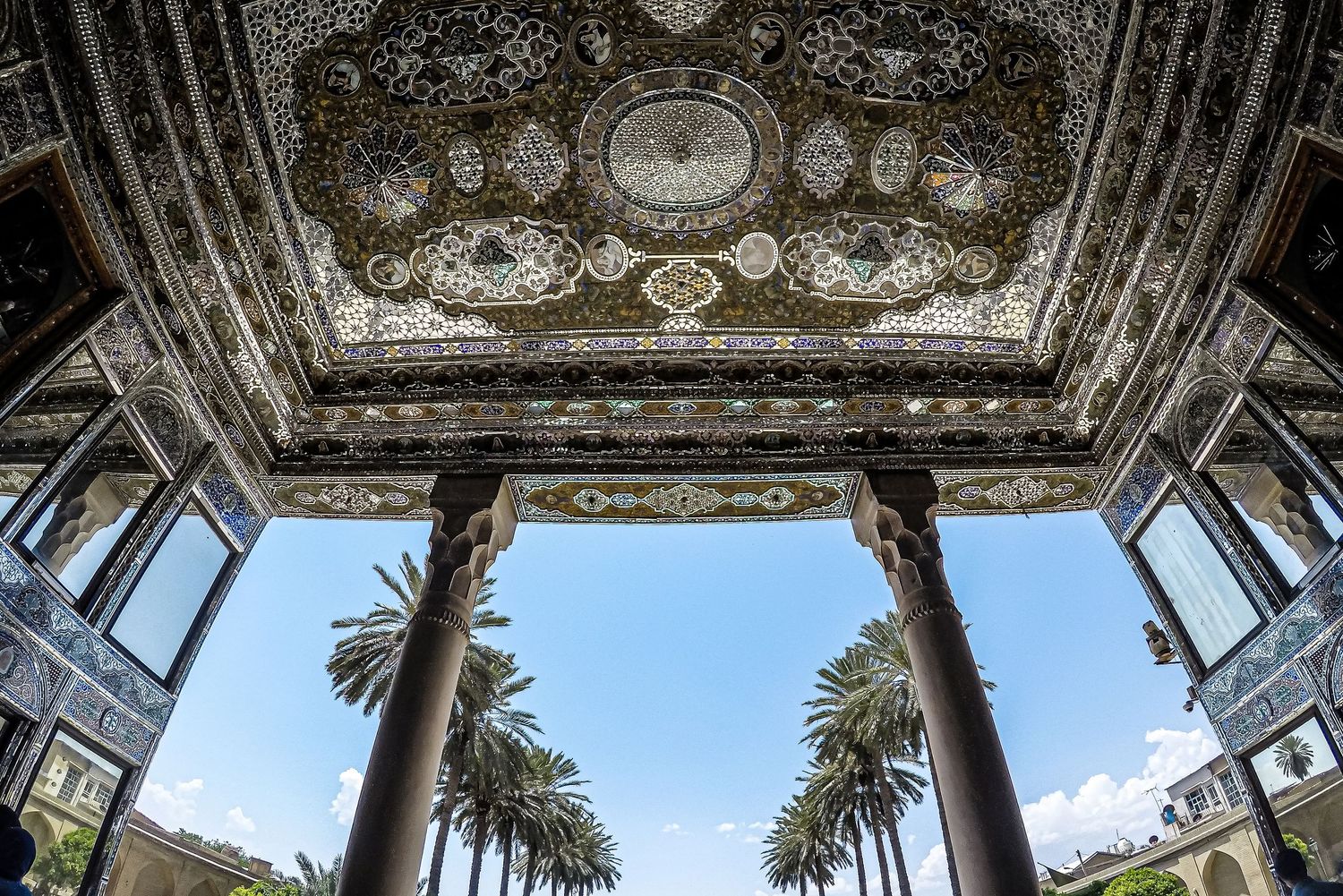 Ornate palace pavilion with mirrored ceiling and columns framing palm trees and sky.