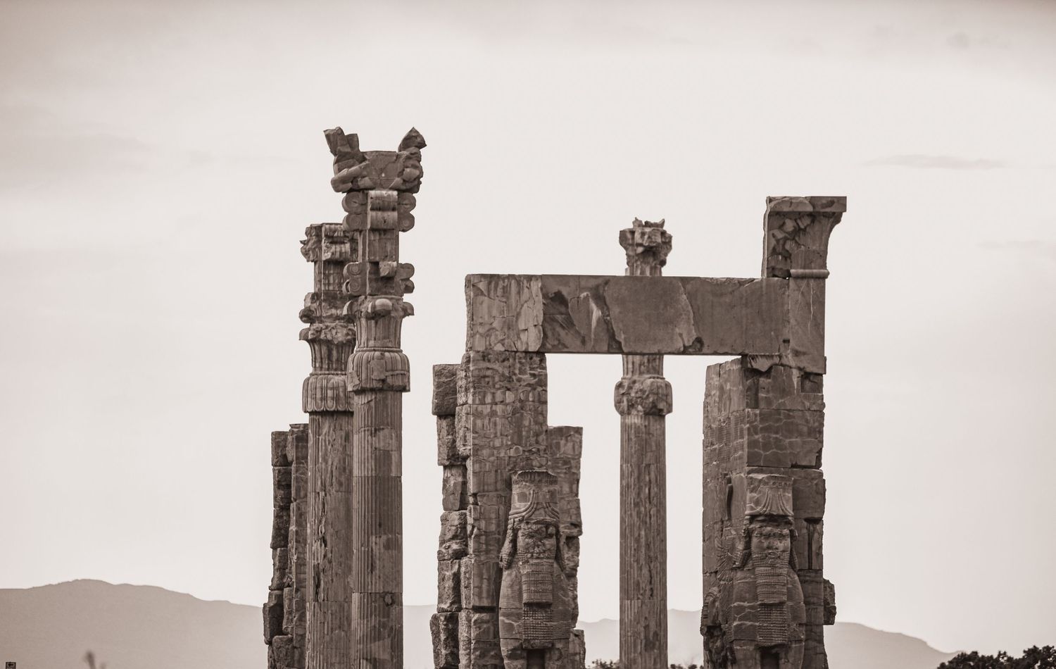 Ancient stone columns and architectural ruins stand against a misty mountain backdrop in sepia tones.