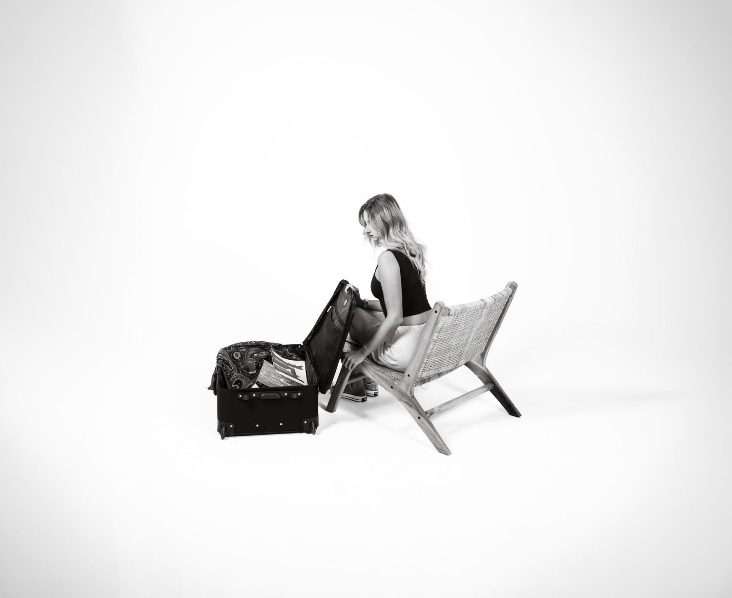 Black and white photo of someone sitting on a chair next to a suitcase in a minimalist setting.