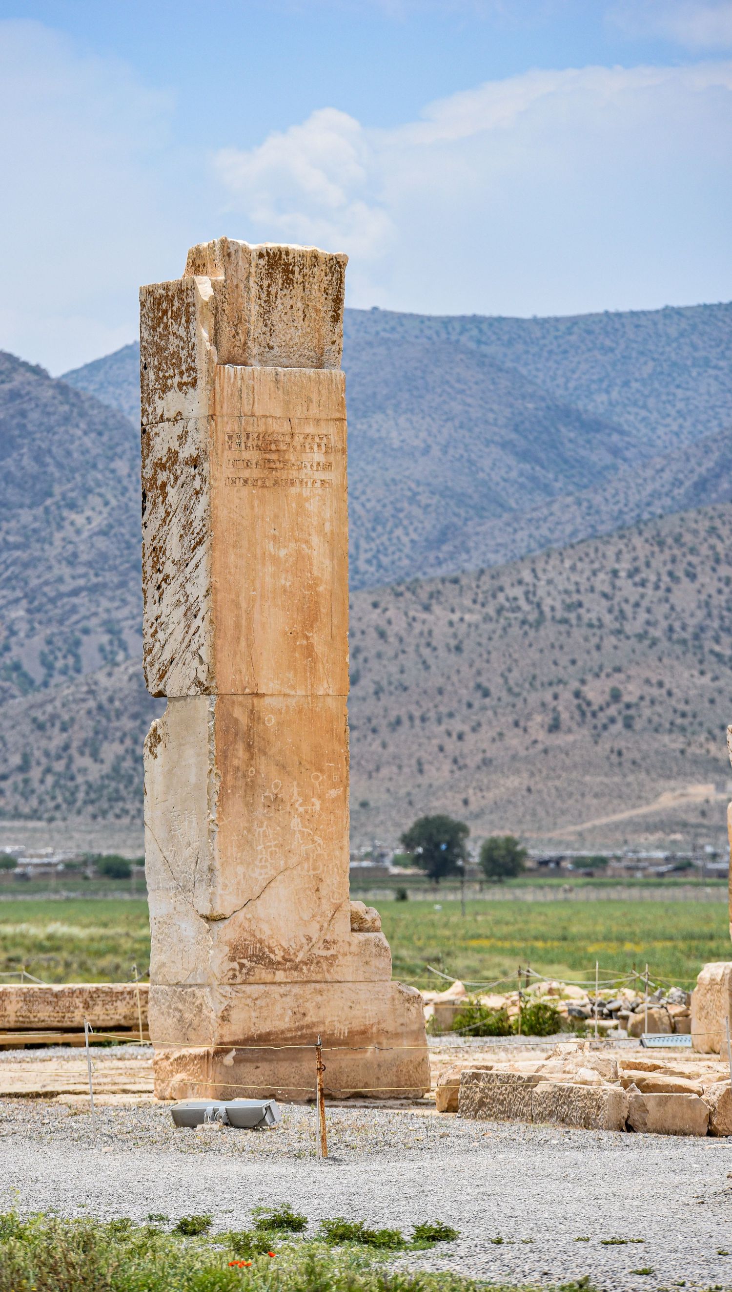 Lone standing stone pillar with inscriptions amid arid landscape with mountains in background.