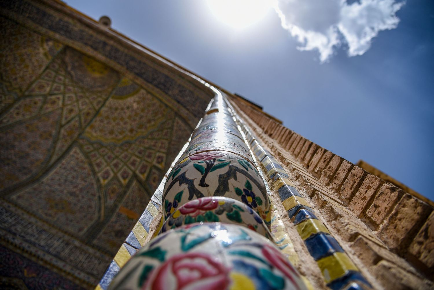 Ornate decorative column with colorful tilework patterns reaches upward toward a bright sun and cloudy sky.