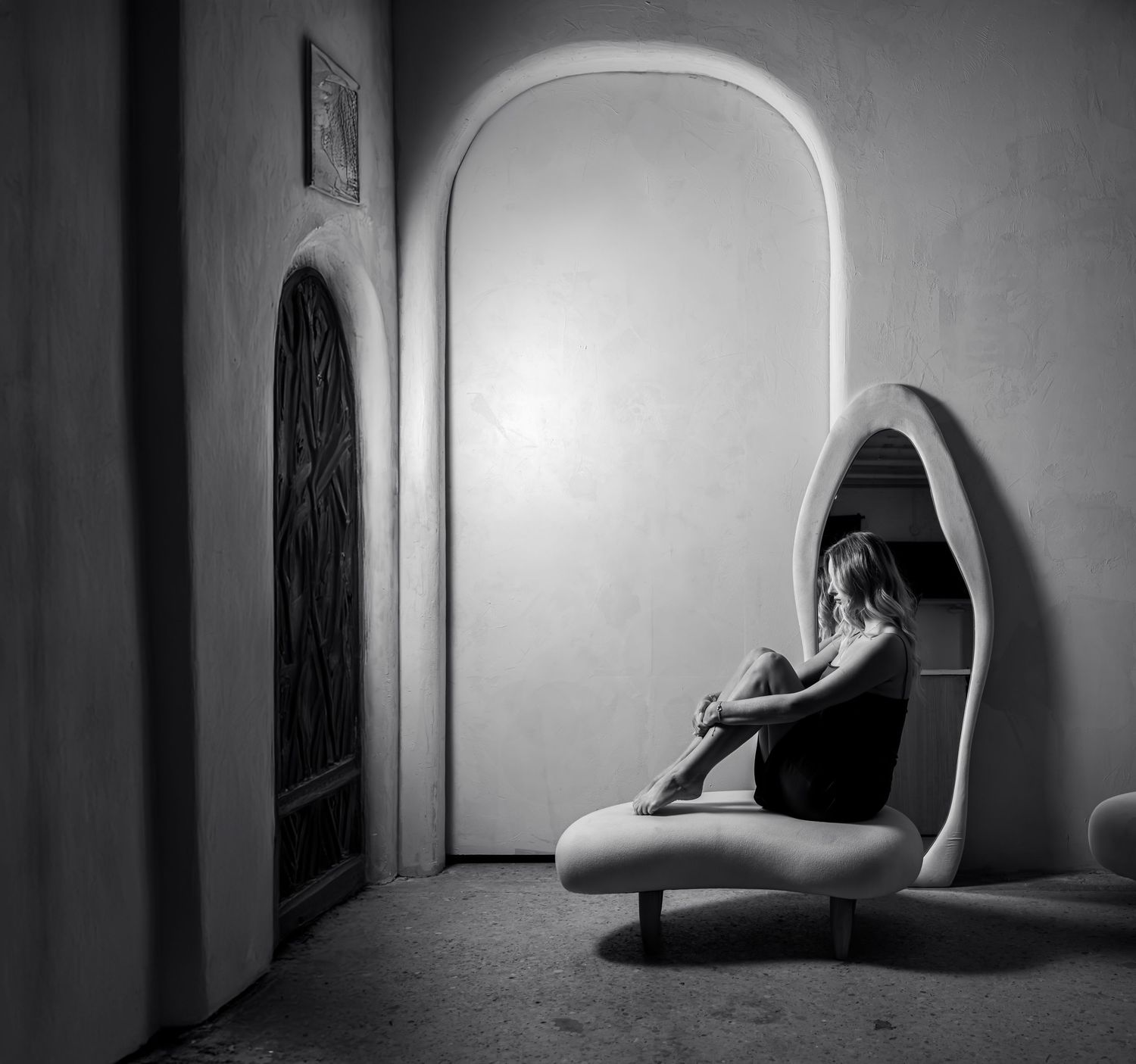 Artistic black and white portrait of person curled up in egg-shaped chair by arched doorway.