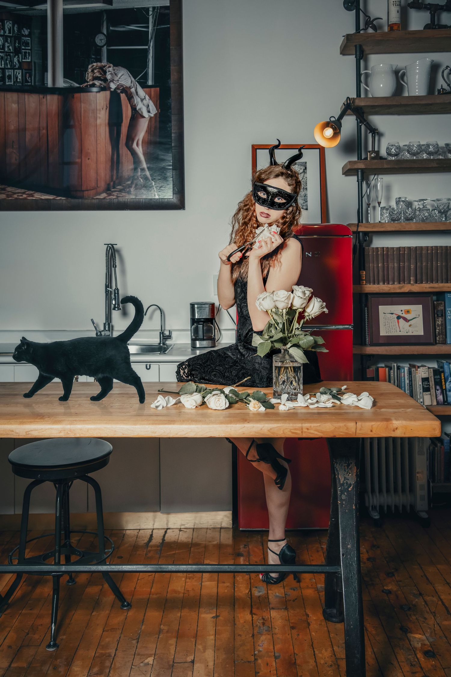 Theatrical scene with black cat walking past masked figure arranging flowers on wooden table.