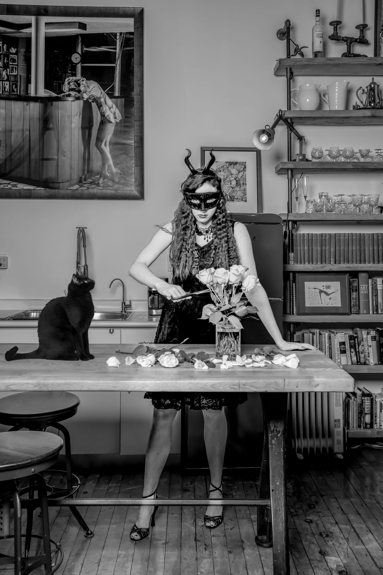 Woman in mask arranging flowers while black cat watches from kitchen counter in black and white photo.