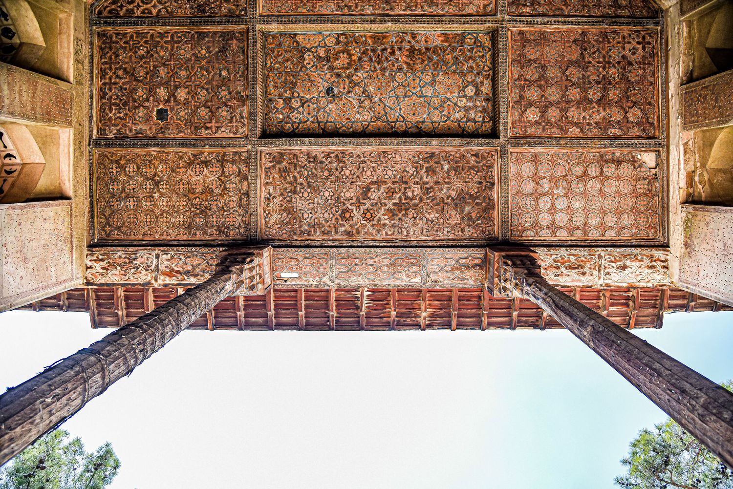 Ancient carved wooden ceiling detail viewed from below, framed by stone columns against blue sky.