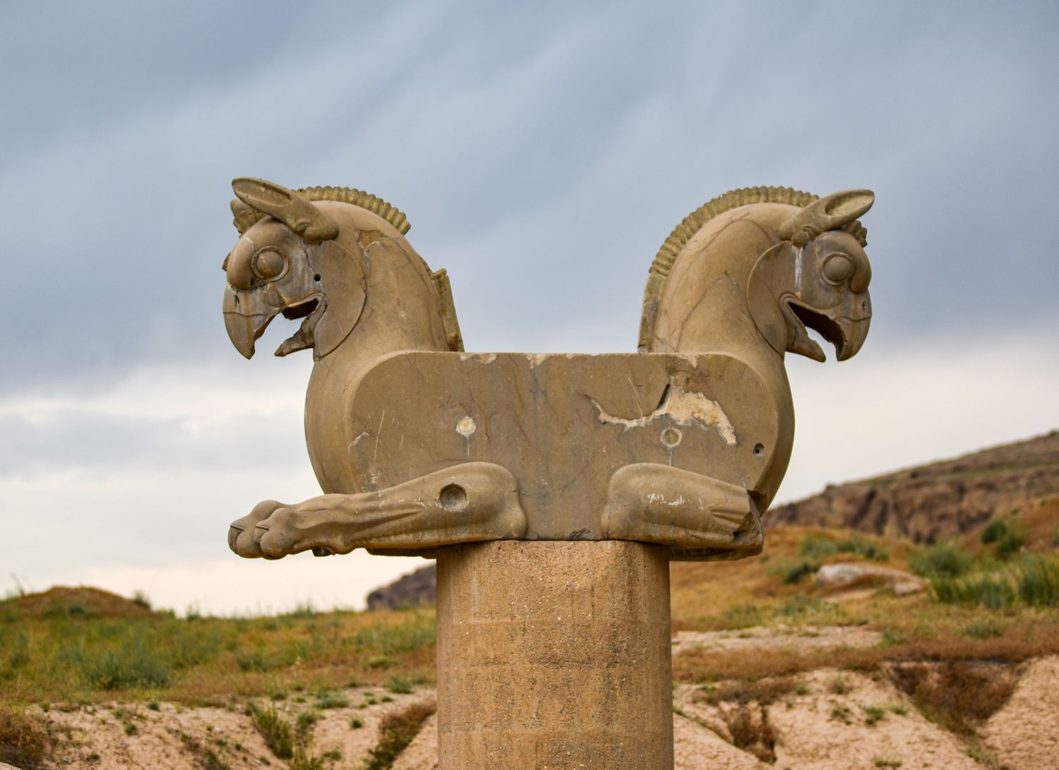 Double-headed horse capital sculpture atop stone column against cloudy sky.