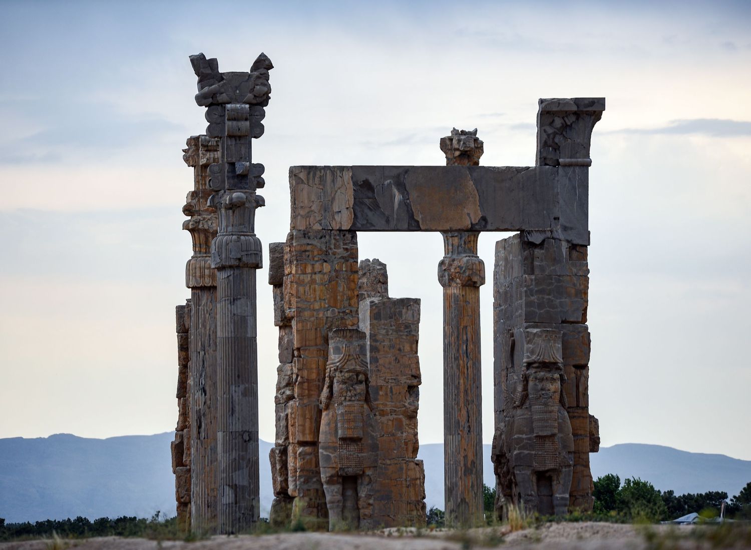 Towering stone pillars of an ancient Persian palace frame mountain views at sunset.
