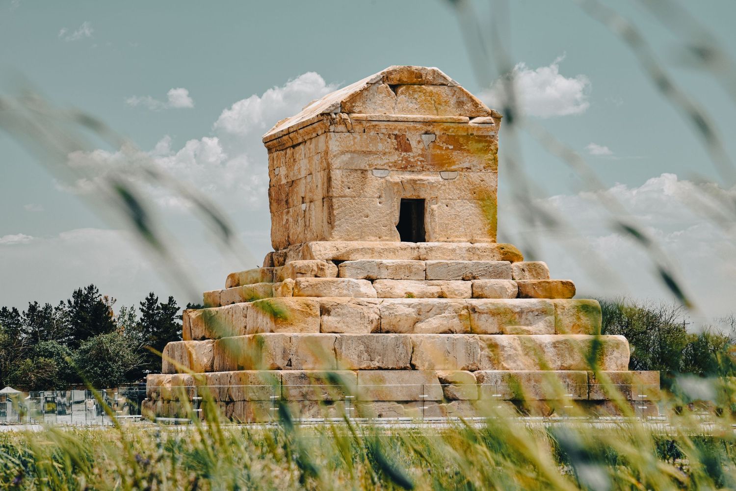 Ancient stone tomb structure with stepped base rises among grass fields against blue sky.