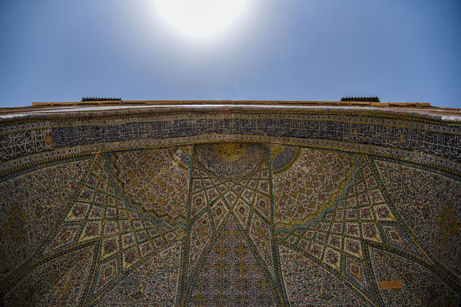 Looking up at ornate Islamic architectural details carved into exterior dome against bright blue sky.