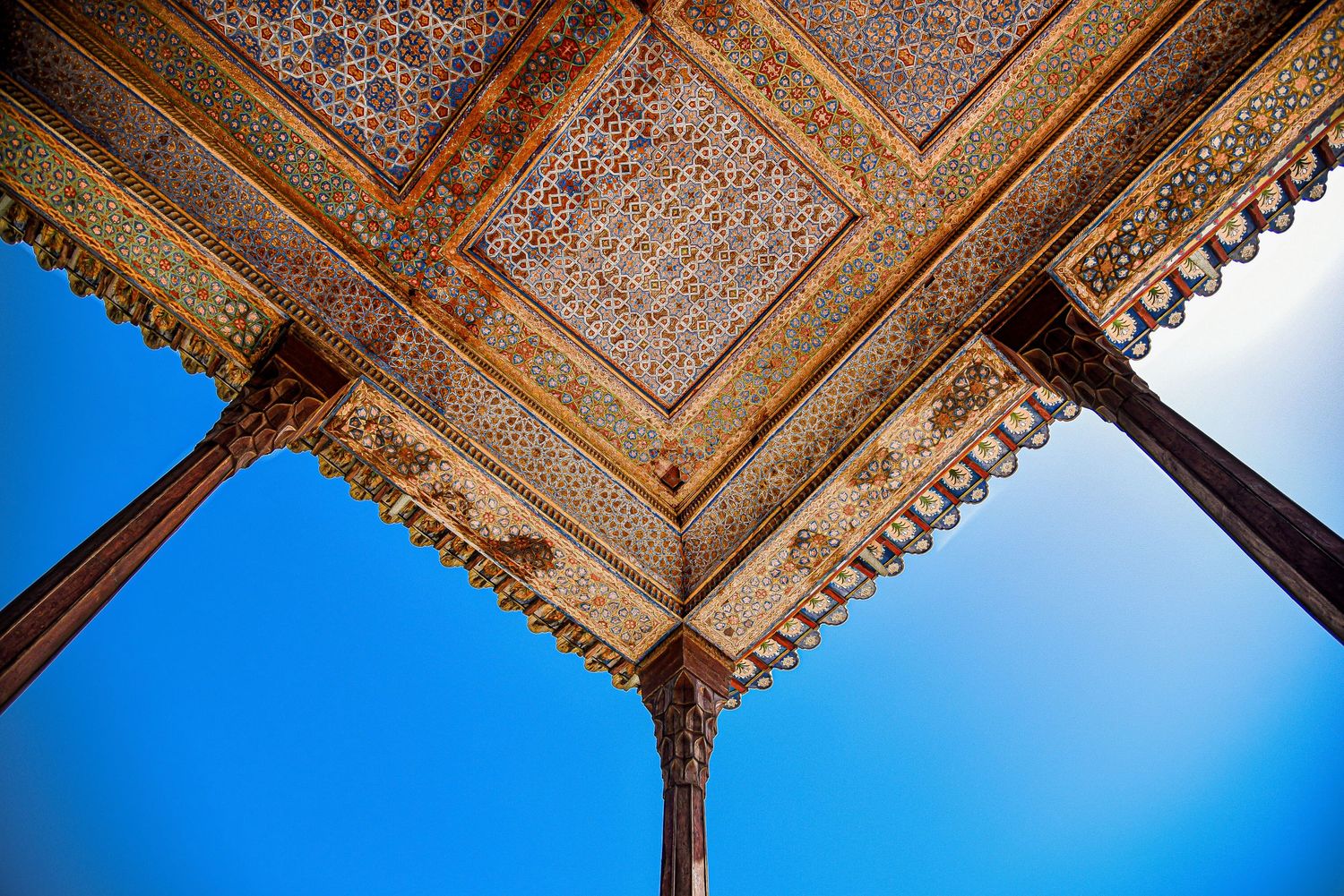 Looking up at an elaborately carved wooden ceiling with geometric patterns against bright blue sky.