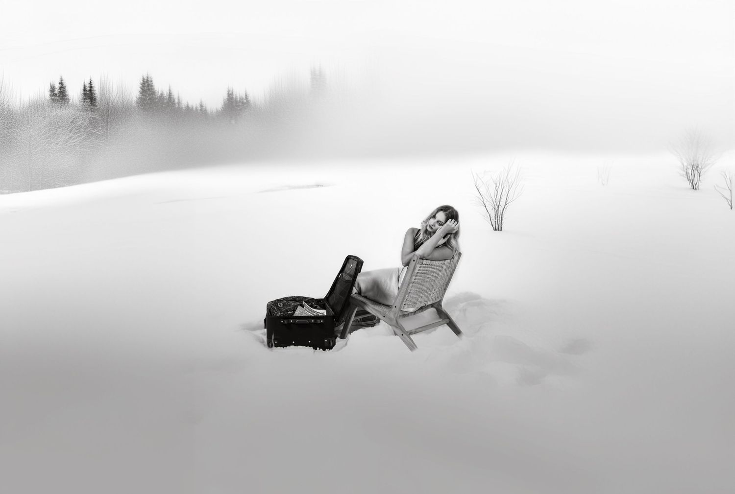 Black suitcase and beach chair sitting in snowy landscape with misty evergreen trees in background.
