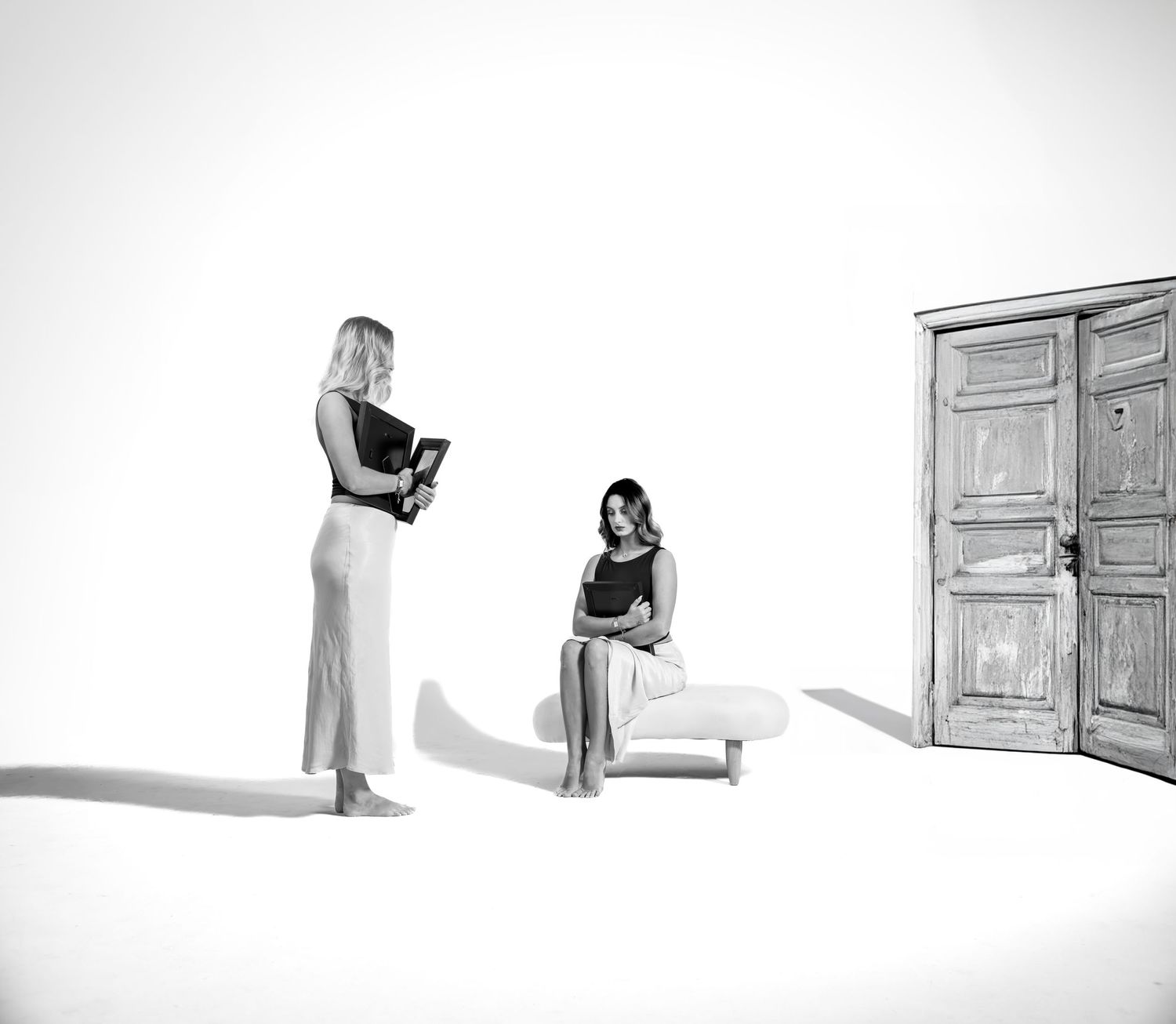 Minimalist black and white photograph of two people in a stark white room with wooden door.