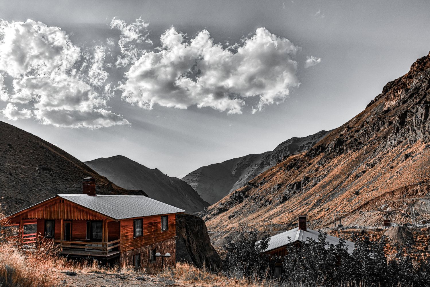 Rustic wooden cabin nestled in mountainous valley with dramatic cloudy sky overhead.