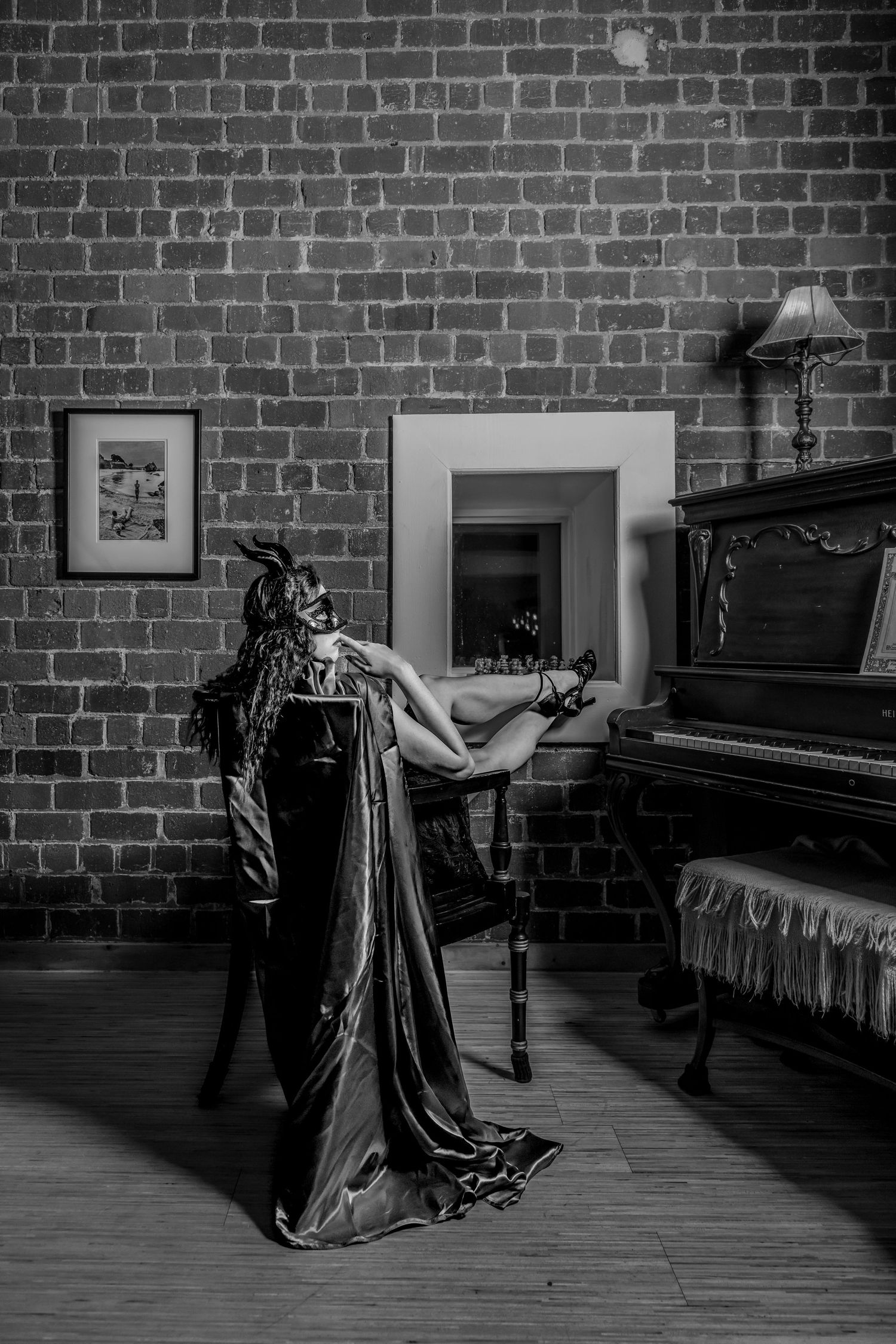 Dramatic black and white photo of masked figure in flowing gown posing near vintage piano.