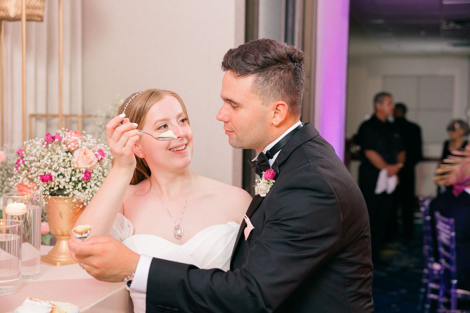 Newlyweds dance together in a beautifully lit ballroom with pink and purple ambient lighting.