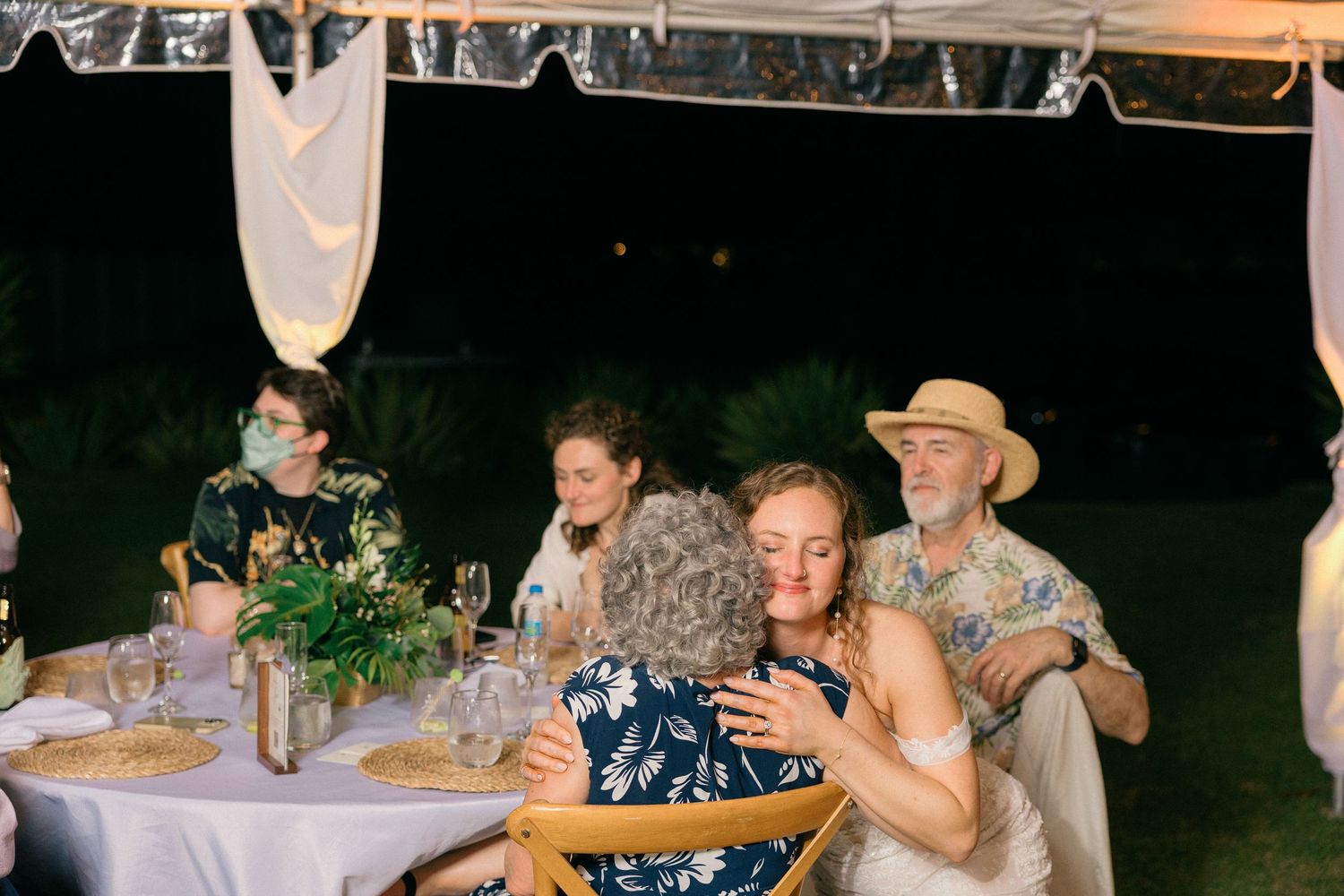Wedding reception guests seated at long tables under a white tent canopy at night.