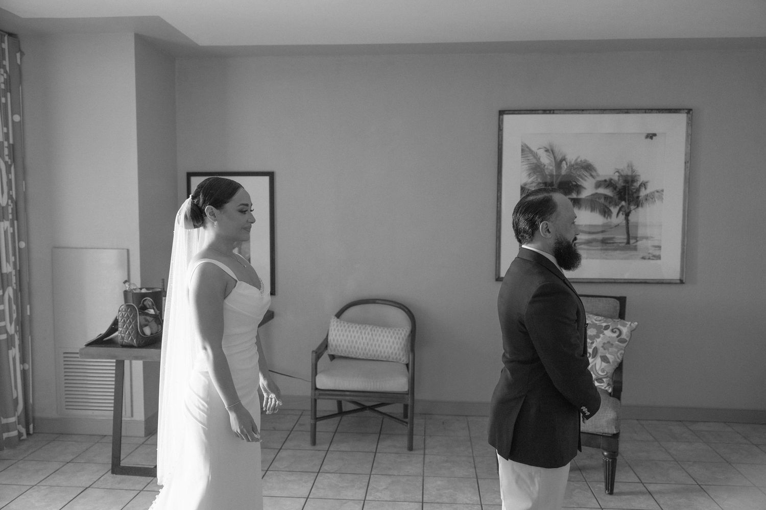 A first look wedding moment as a bride in a sleek white dress stands behind her waiting partner.
