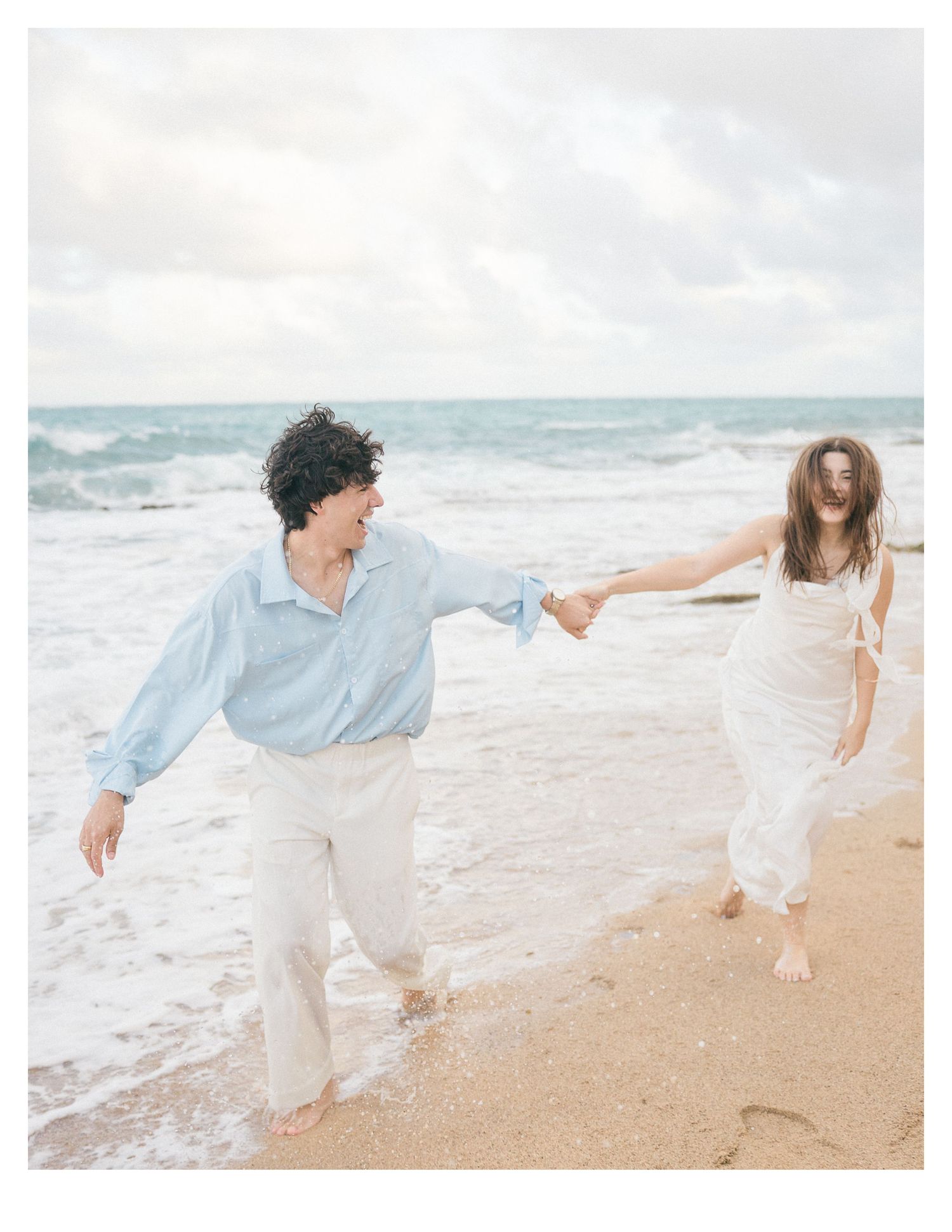 Couple in light clothing holding hands while running on beach at sunset.