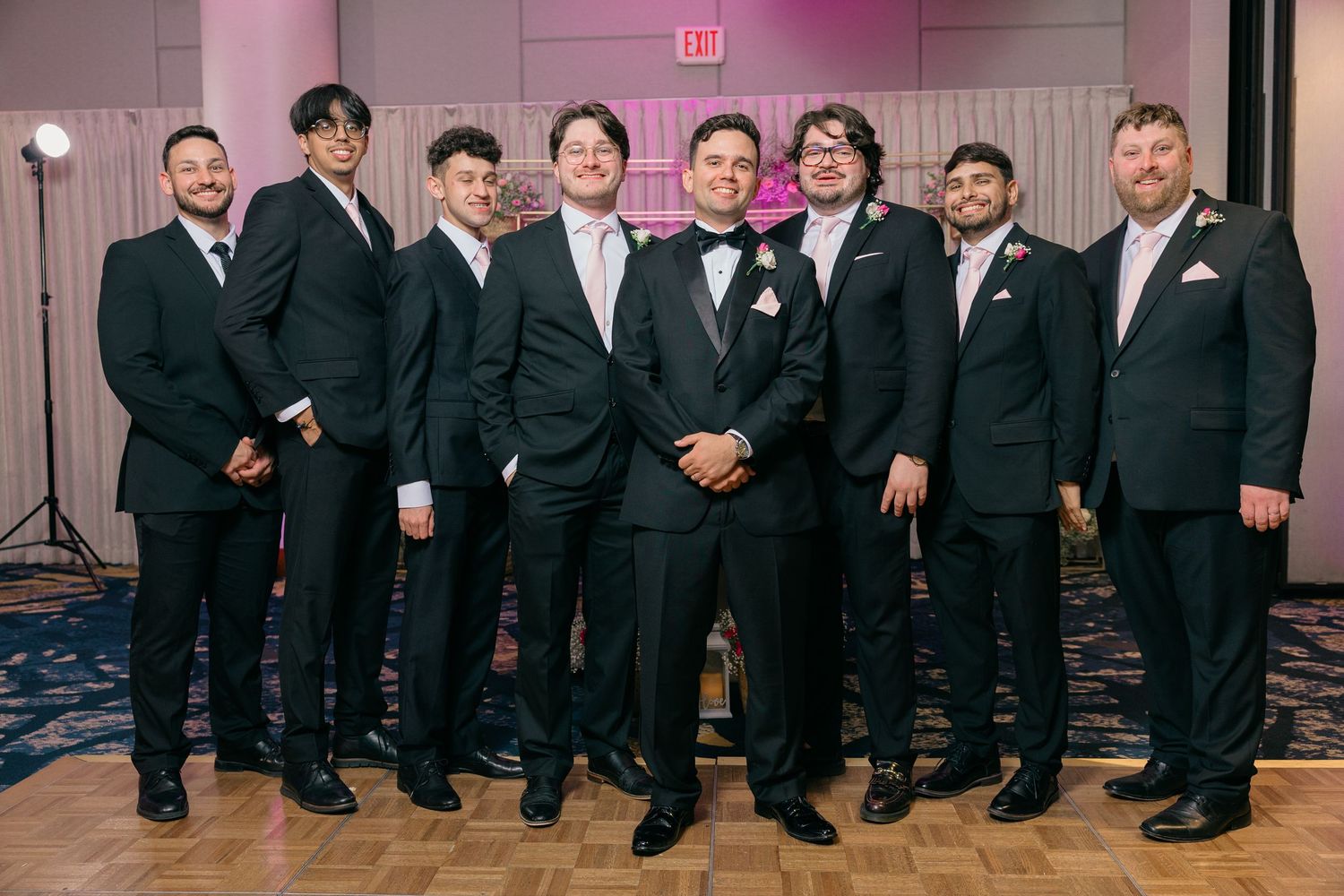 Formal wedding party lined up in black suits against pink-lit backdrop.