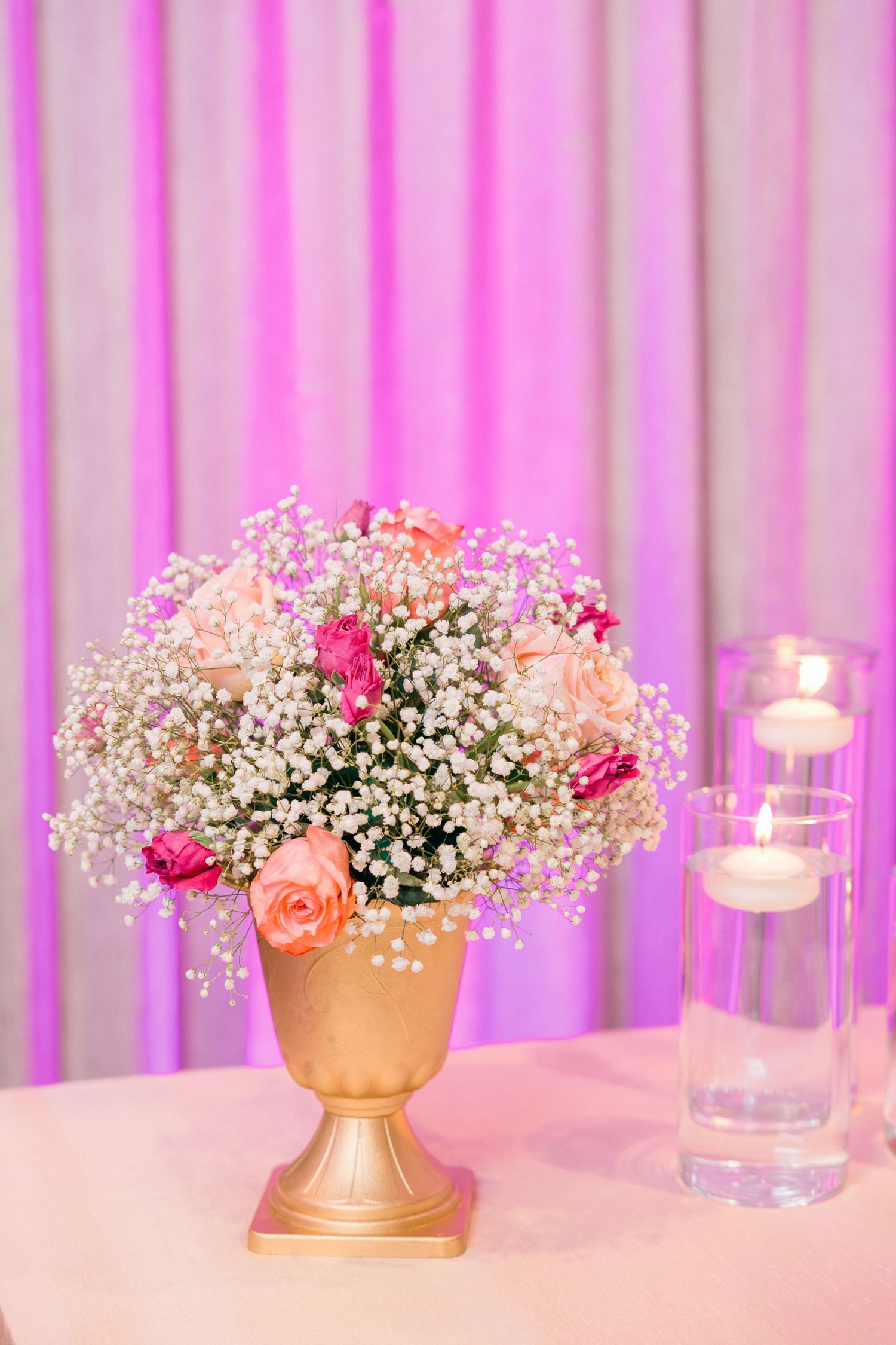 Elegant floral centerpieces with pink and peach roses and baby's breath in gold vases against a pink-lit backdrop.