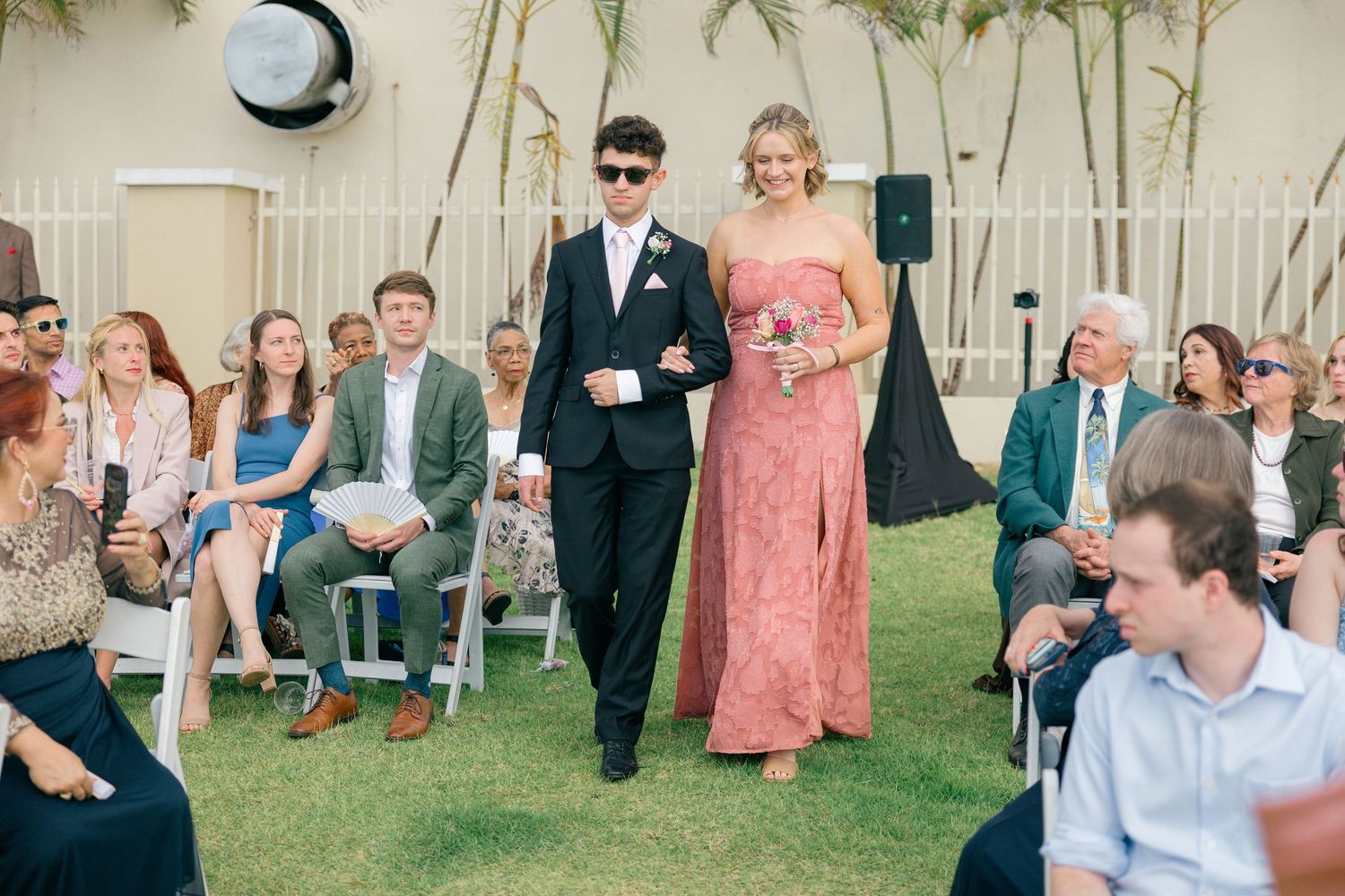 Wedding guests watching ceremony proceedings on a sunny day with palm trees in the background.