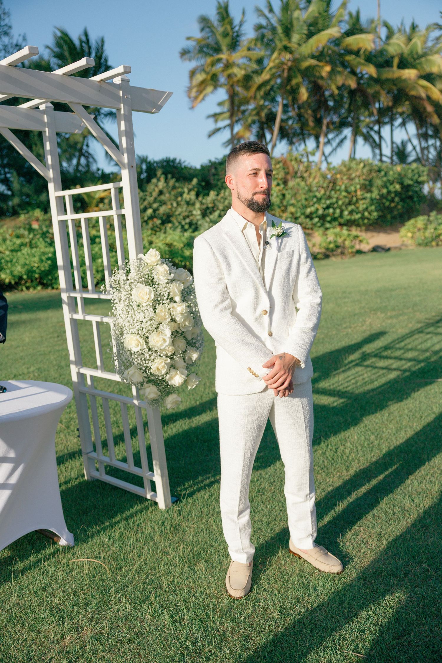 A wedding ceremony setup on a lush green lawn with white decor and palm trees in the tropical background.