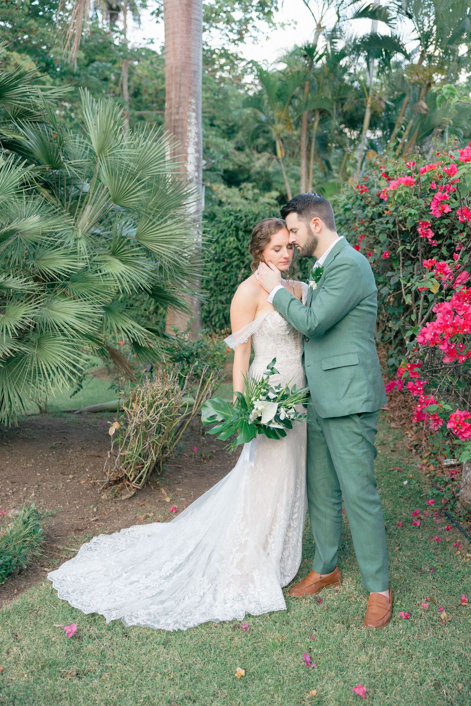 Wedding couple shares intimate moments among tropical landscaping with red bougainvillea flowers.
