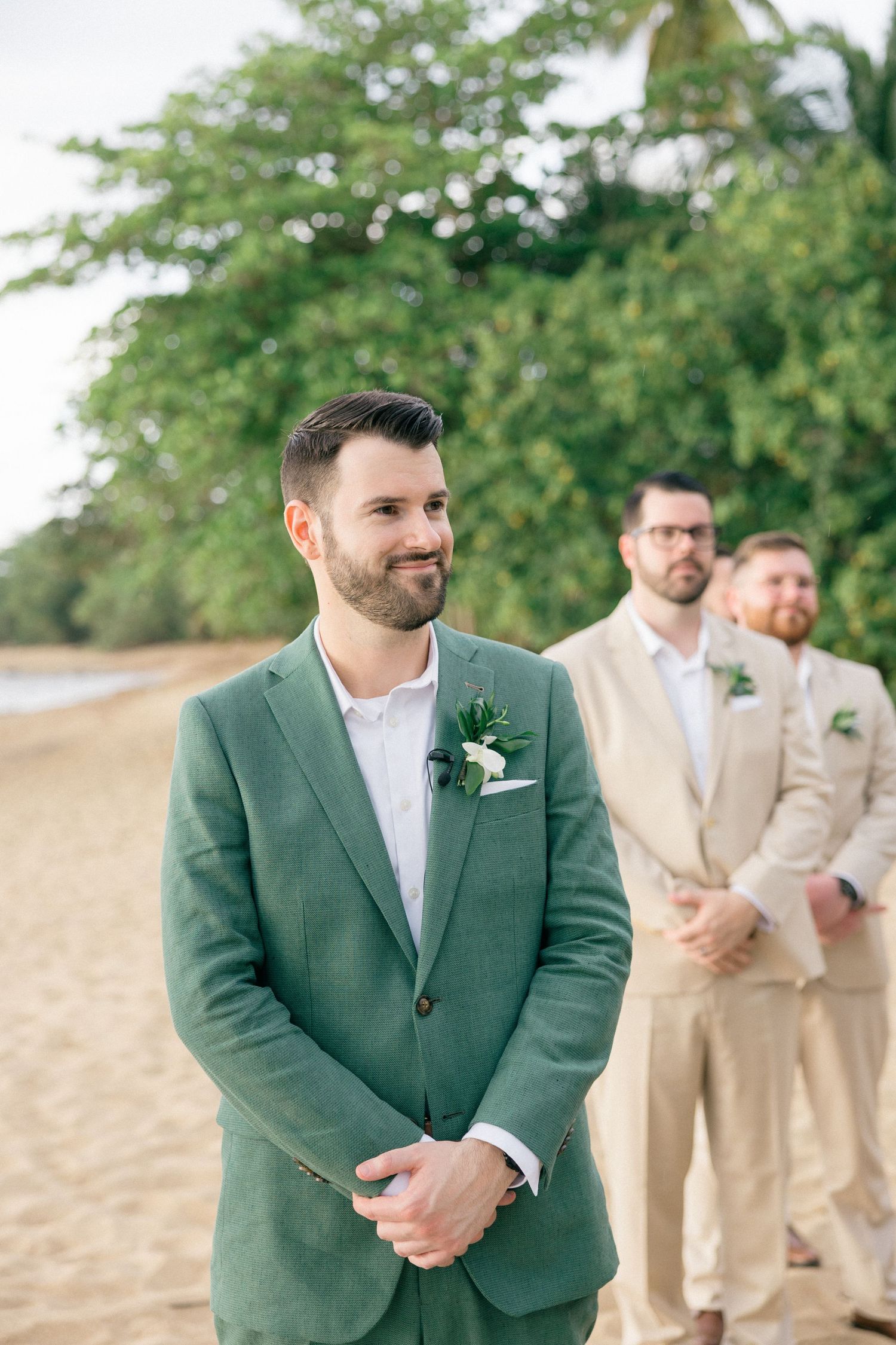 Wedding party members in beige suits and green jackets stand on a beach during a ceremony.