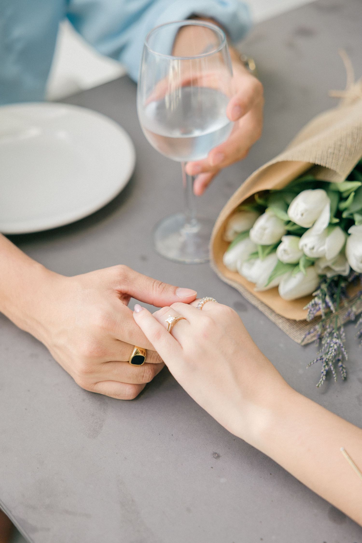 Close-up of hands with engagement ring next to white tulips on a gray surface.