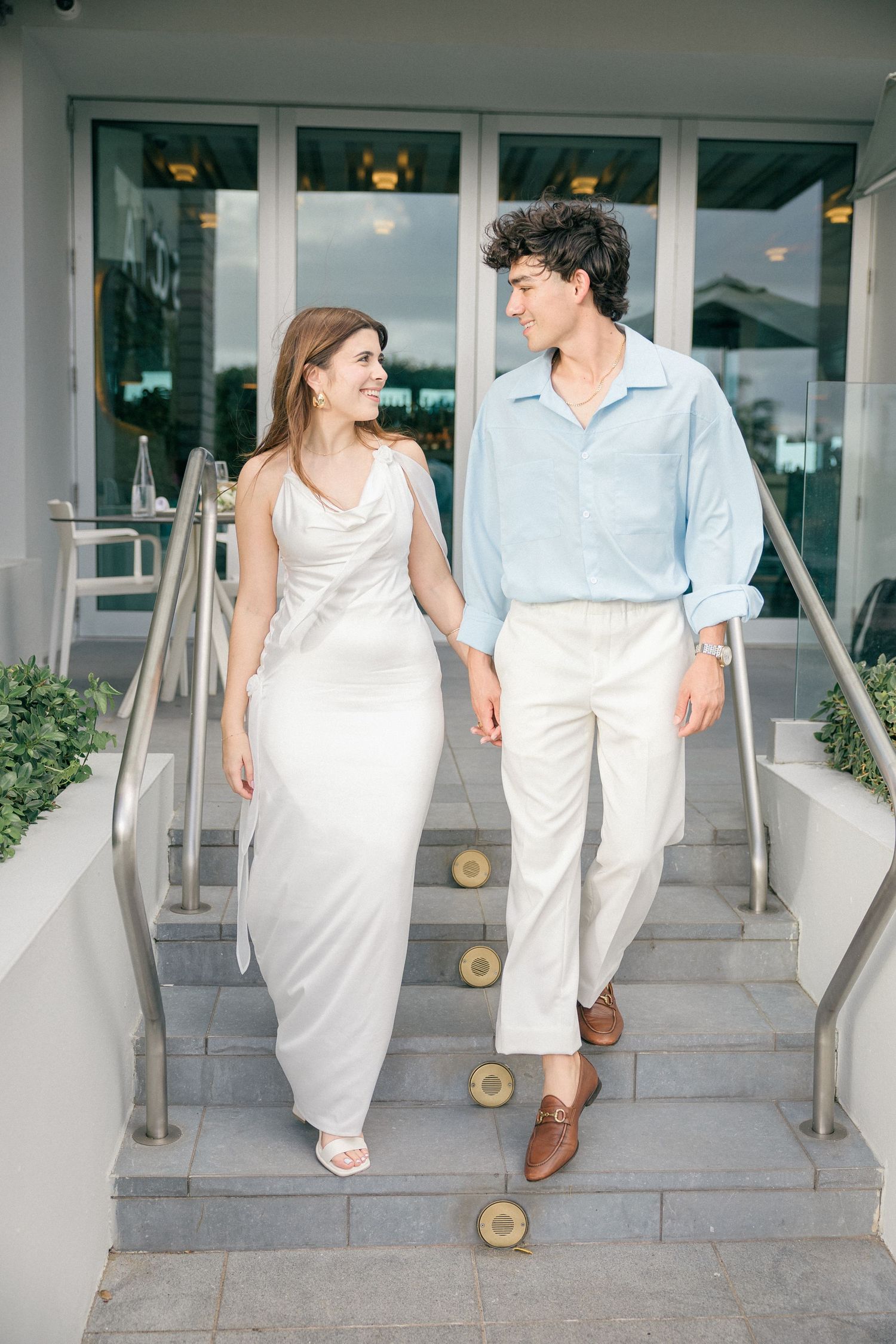 A couple in elegant white and light blue attire walking down outdoor steps together.