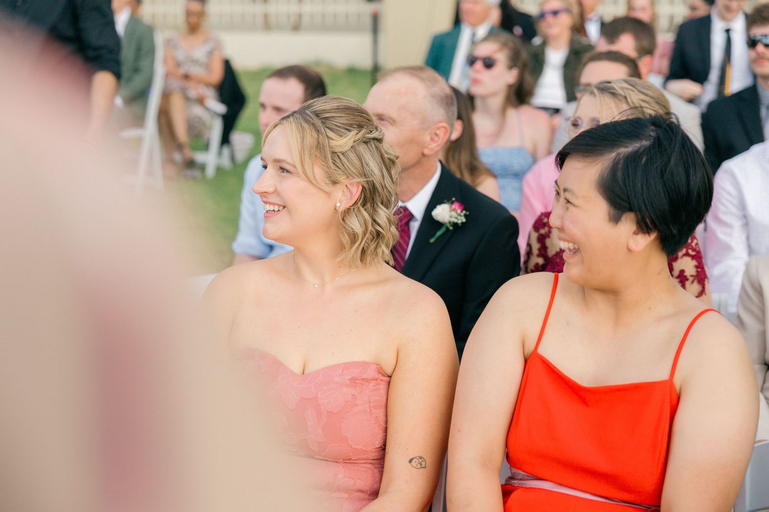 Wedding ceremony attendees in coral and peach dresses seated in outdoor venue.