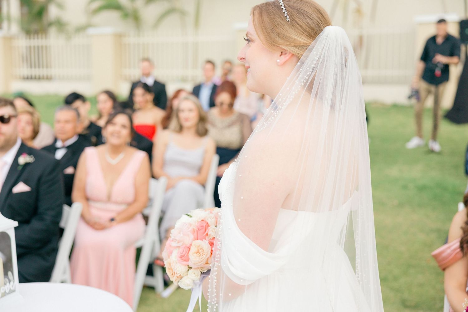 Wedding ceremony guests seated in white chairs on a lawn with palm trees in the background.