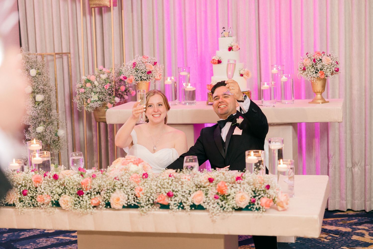 Newlyweds seated at elegant head table with pink lighting and floral decorations.