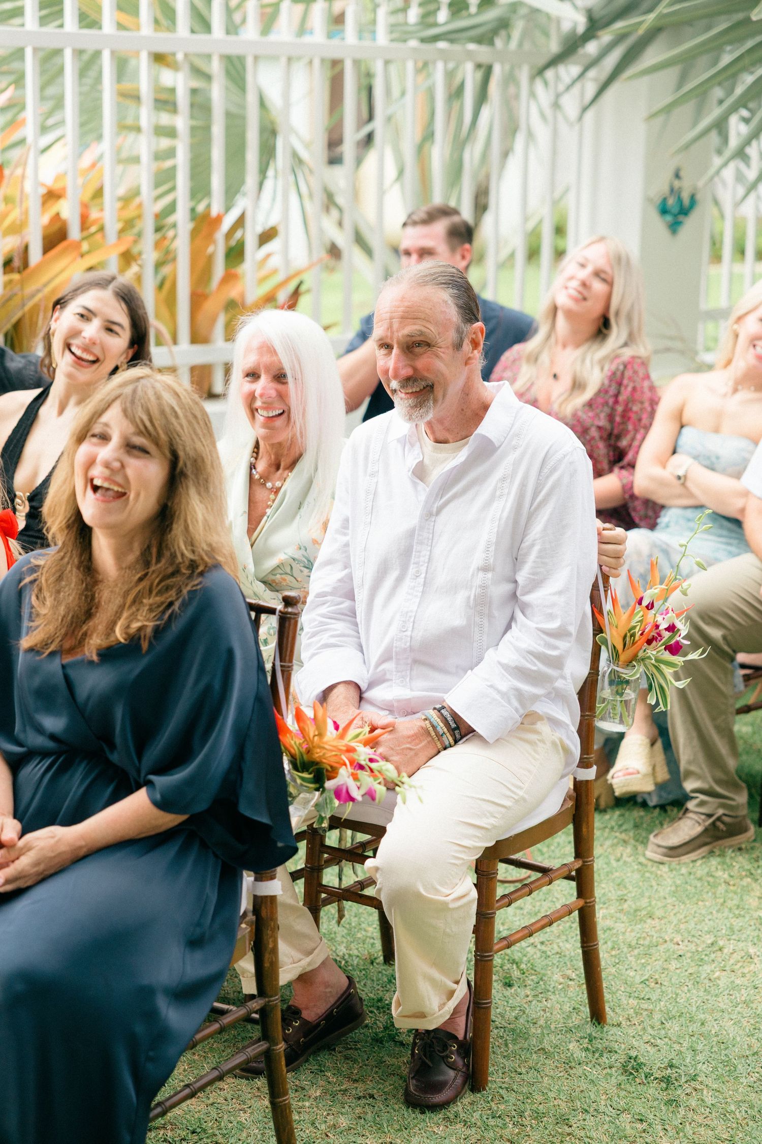 Wedding guests laugh together during outdoor ceremony with tropical greenery backdrop.