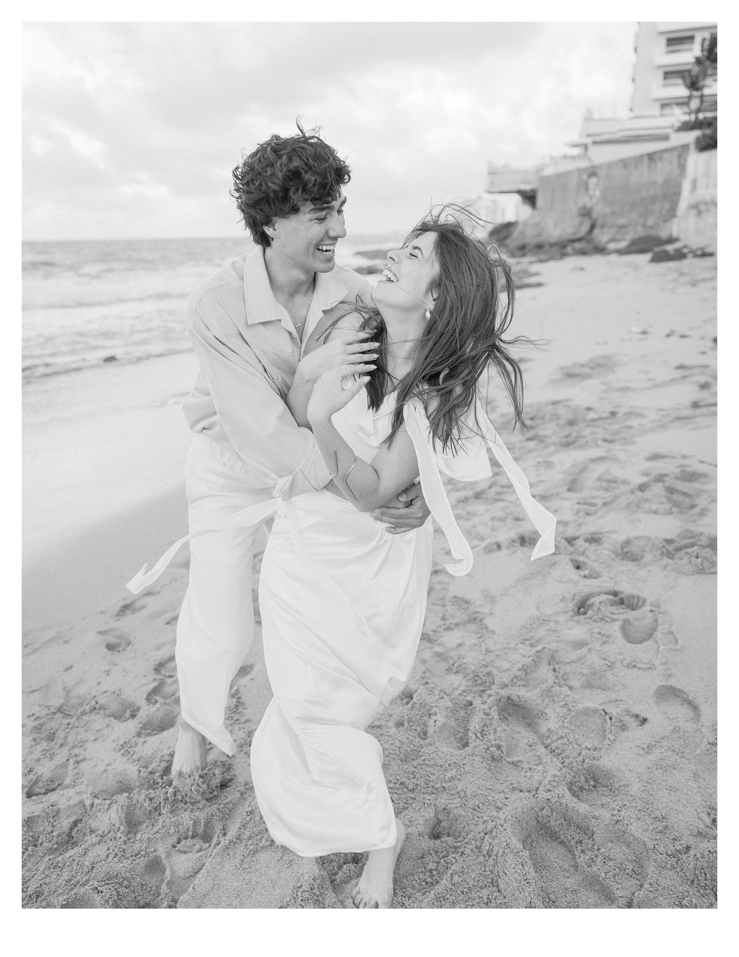 Black and white beach photo of couple laughing and embracing on sandy shore.