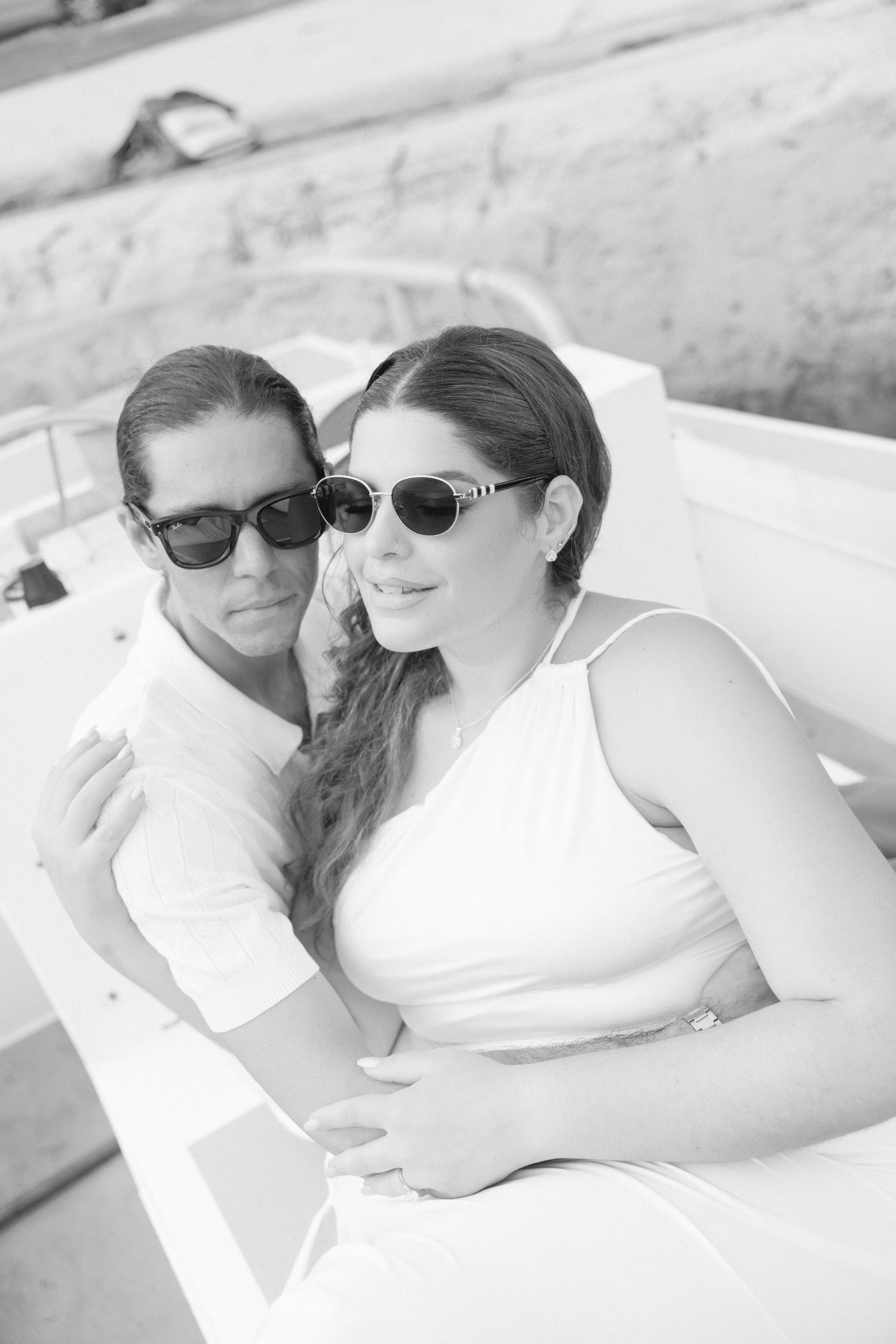 A couple wearing sunglasses relaxes together on white cushions in black and white photography.