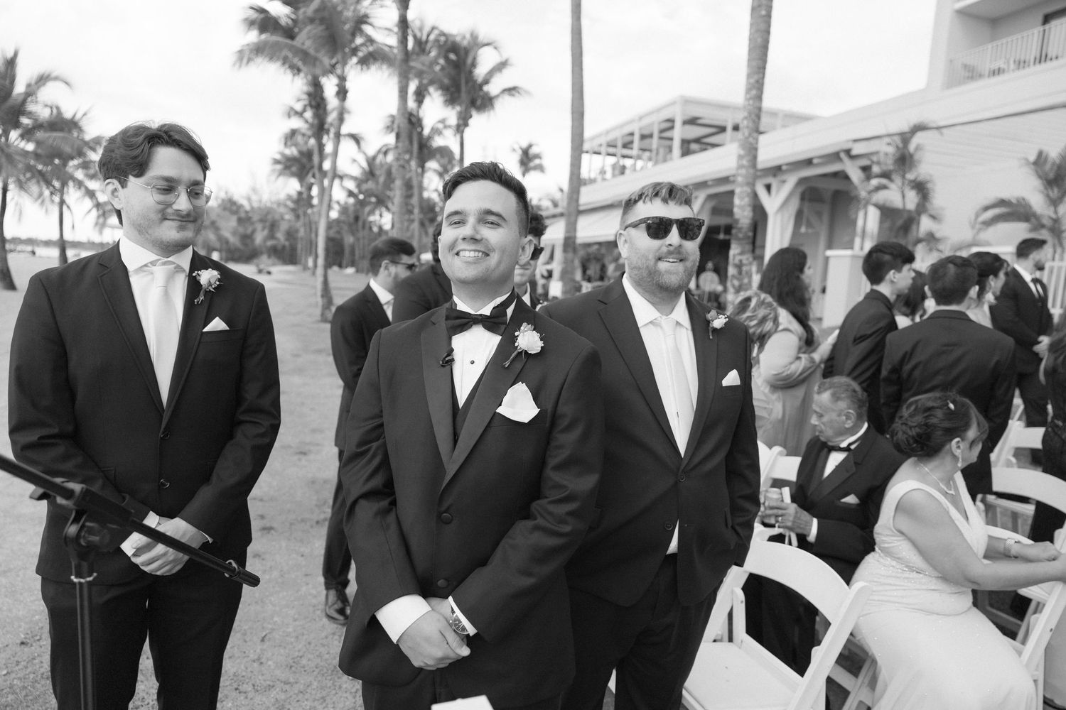 Wedding party members gather outdoors with palm trees and building visible in background.
