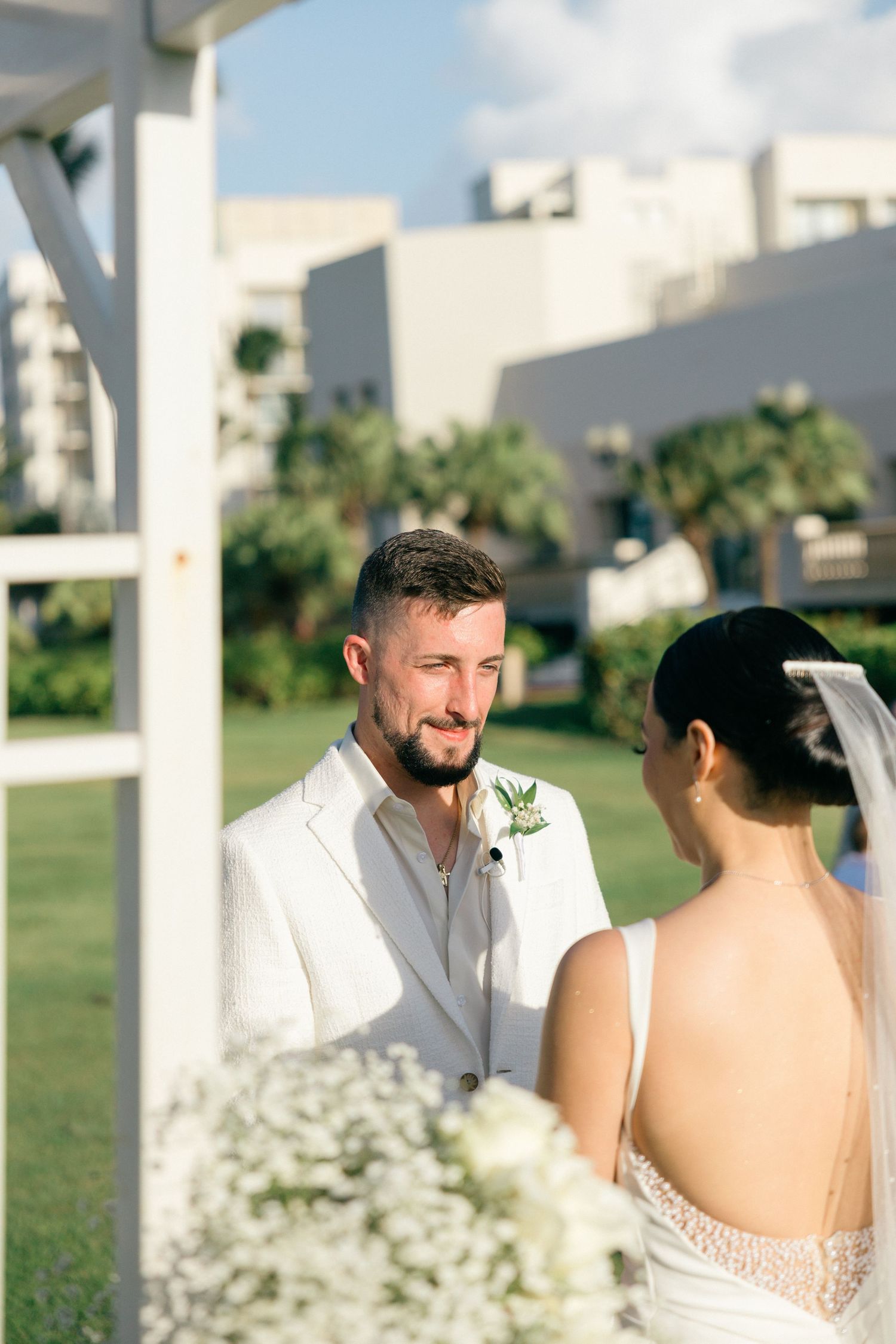 Couple exchanges vows under a white pergola during outdoor ceremony.