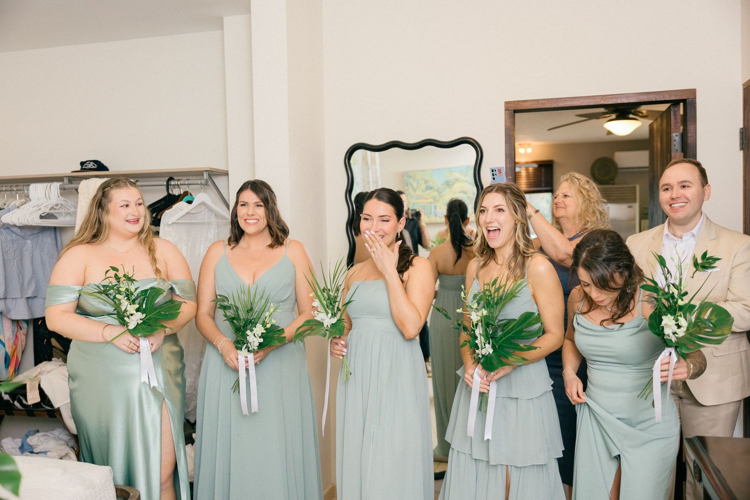Bridesmaids in soft mint green dresses hold lush greenery bouquets while getting ready in a dressing room.