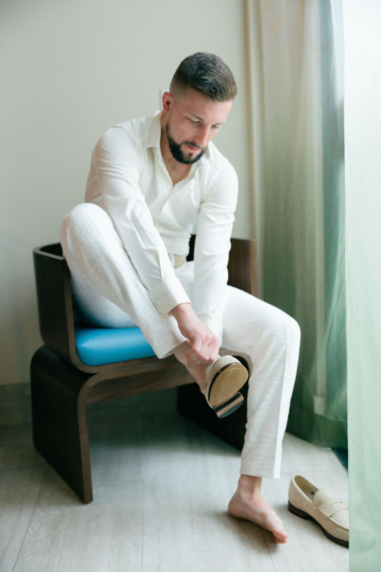 Man in white suit sits contemplatively in a chair by a window.