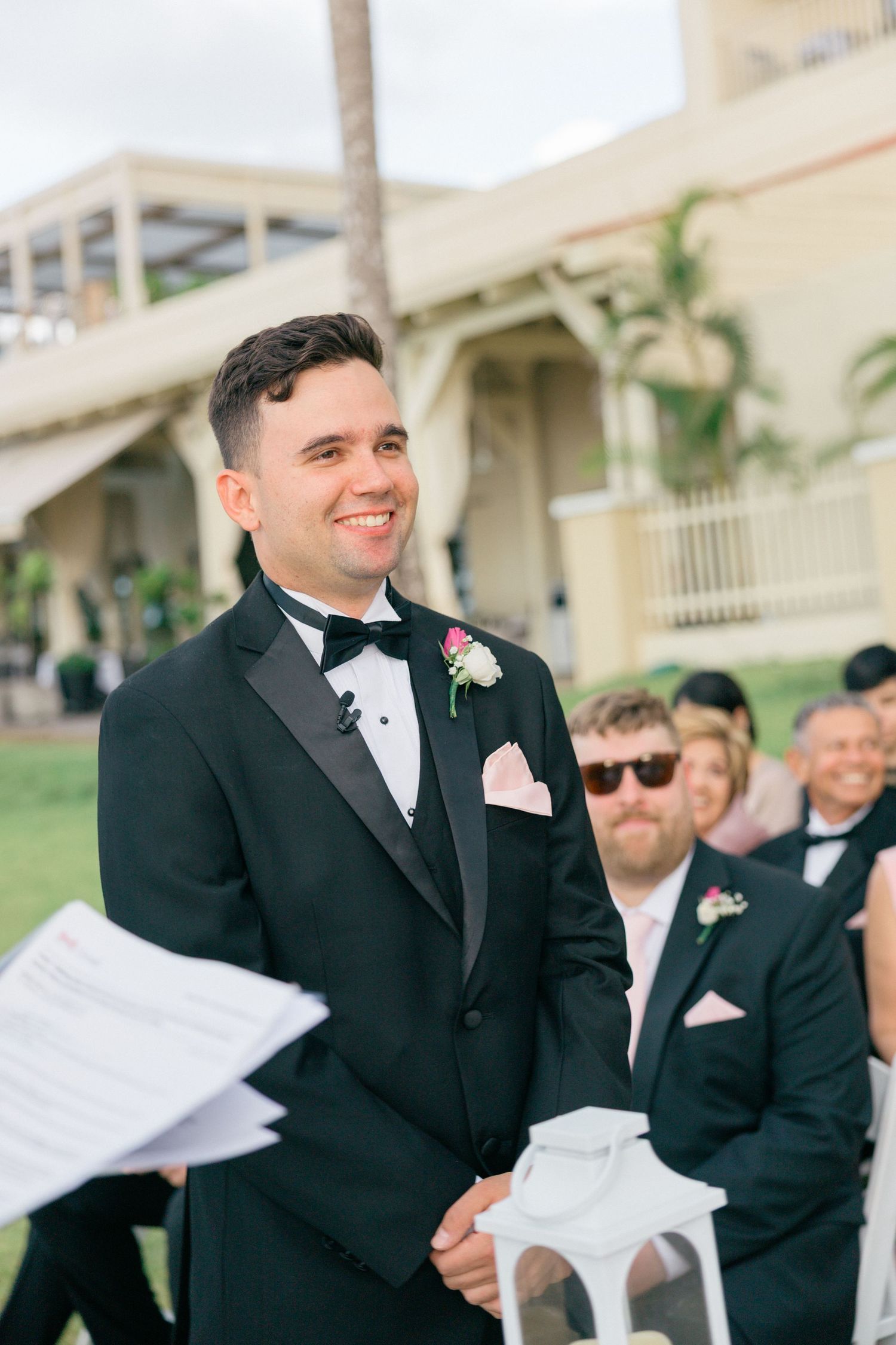 Groomsmen in black tuxedos holding ceremony programs at outdoor wedding.
