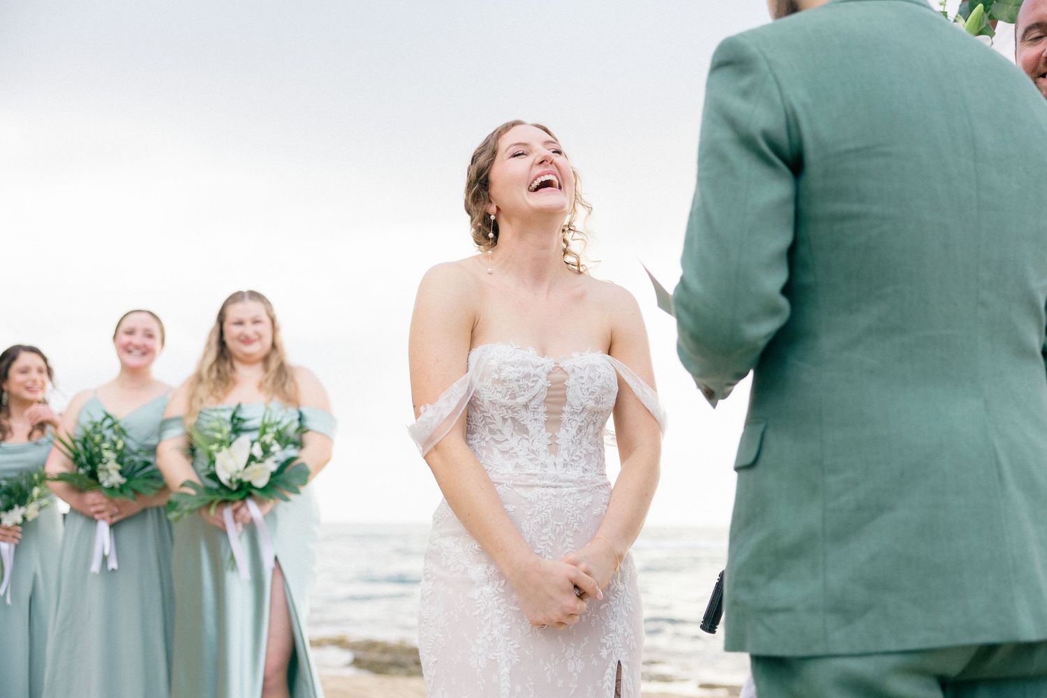 Joyful wedding ceremony on a beach with bridesmaids in sage green dresses and white bouquets.