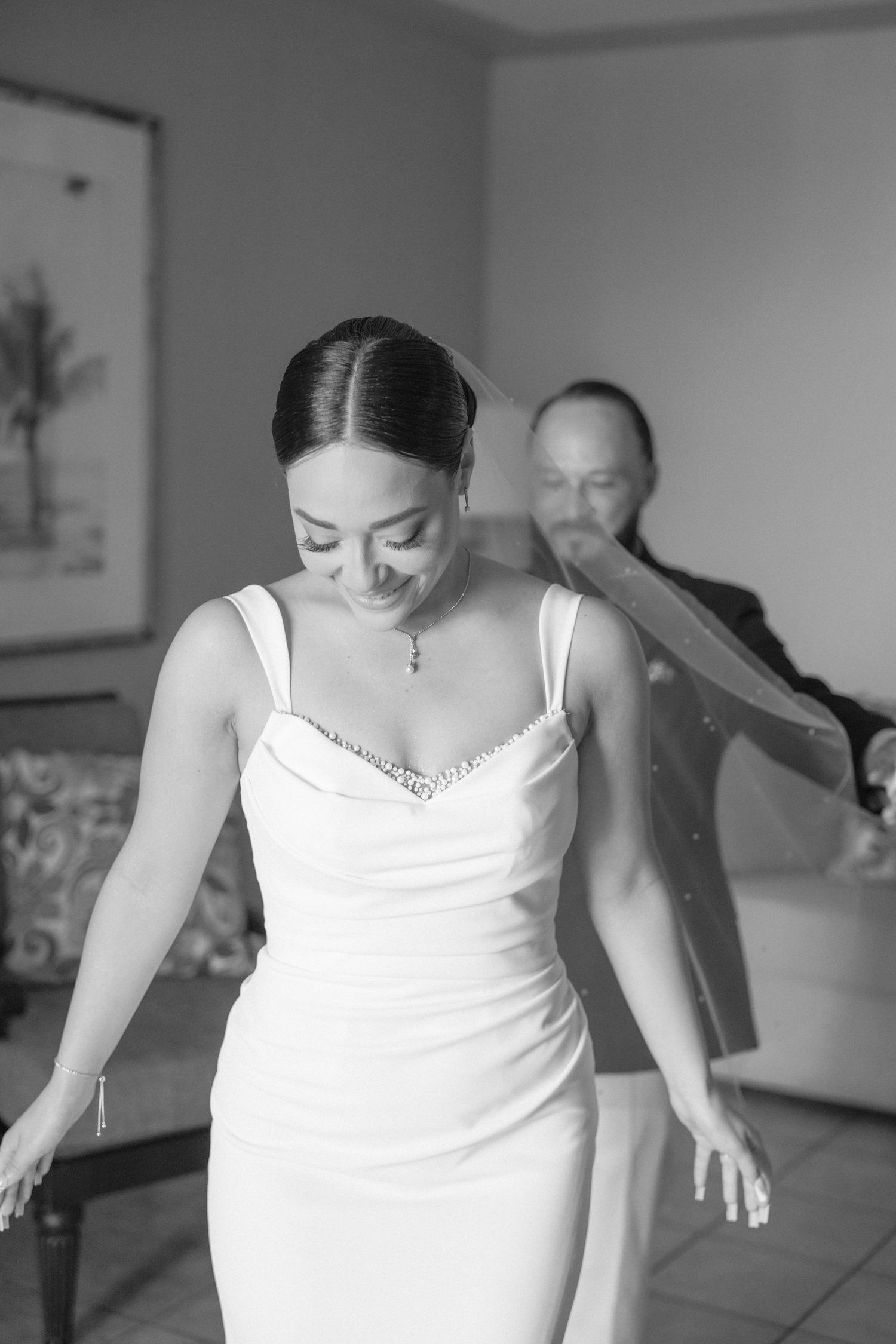 A bride in an elegant white dress smiles while getting ready in a hotel room.