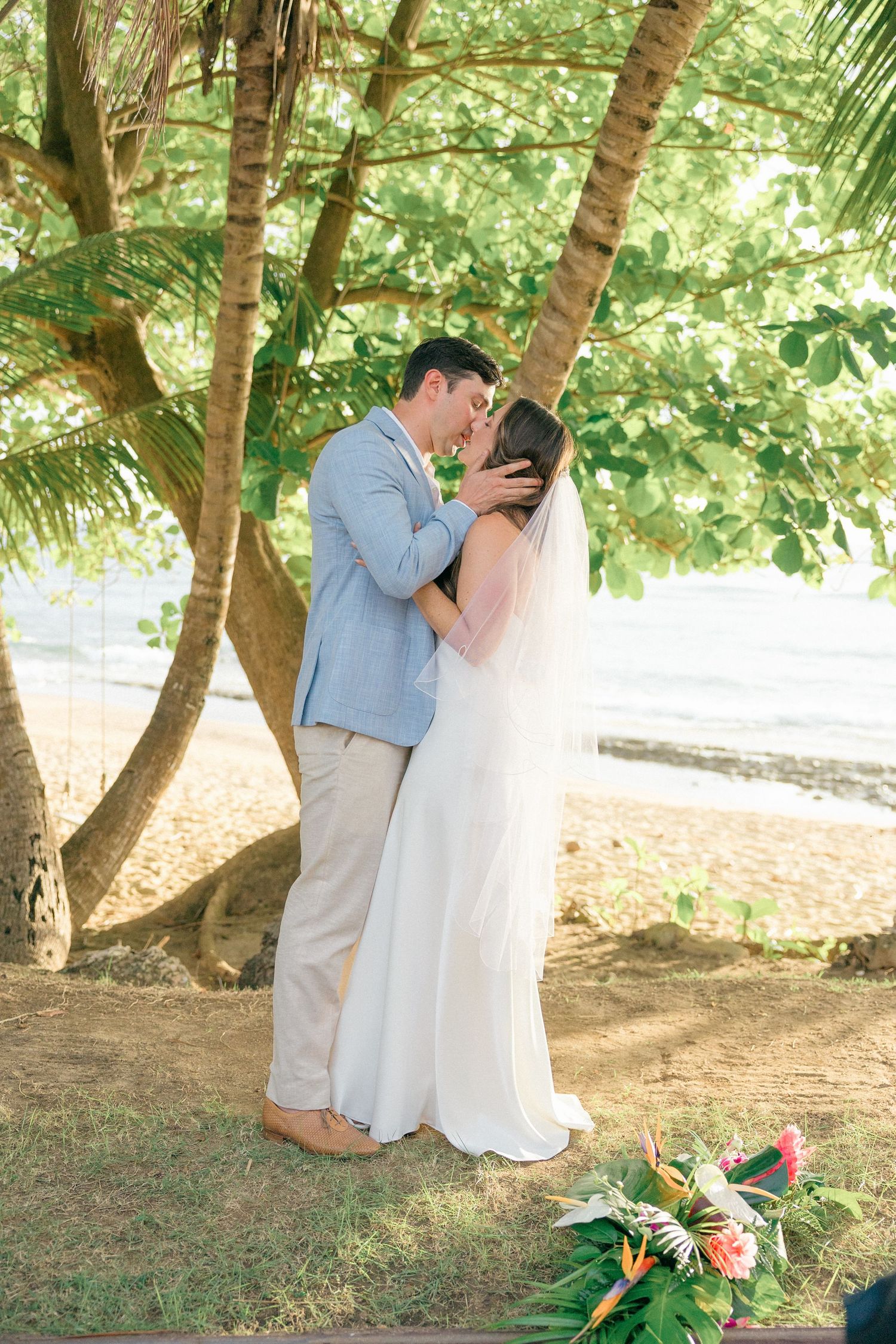 Romantic wedding ceremony moment under tropical trees with floral decorations.